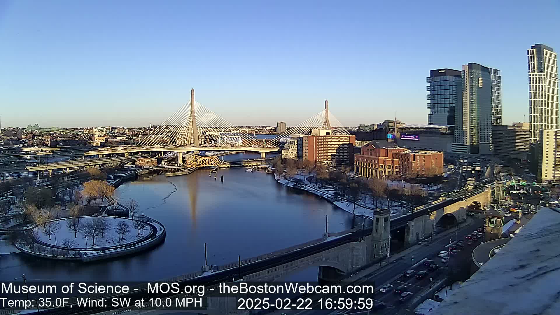 A sunny, cold day with light snow on the ground and a southwest wind shows a city waterfront with two cable-stayed bridges, several high-rise buildings, and a river partially covered with ice.