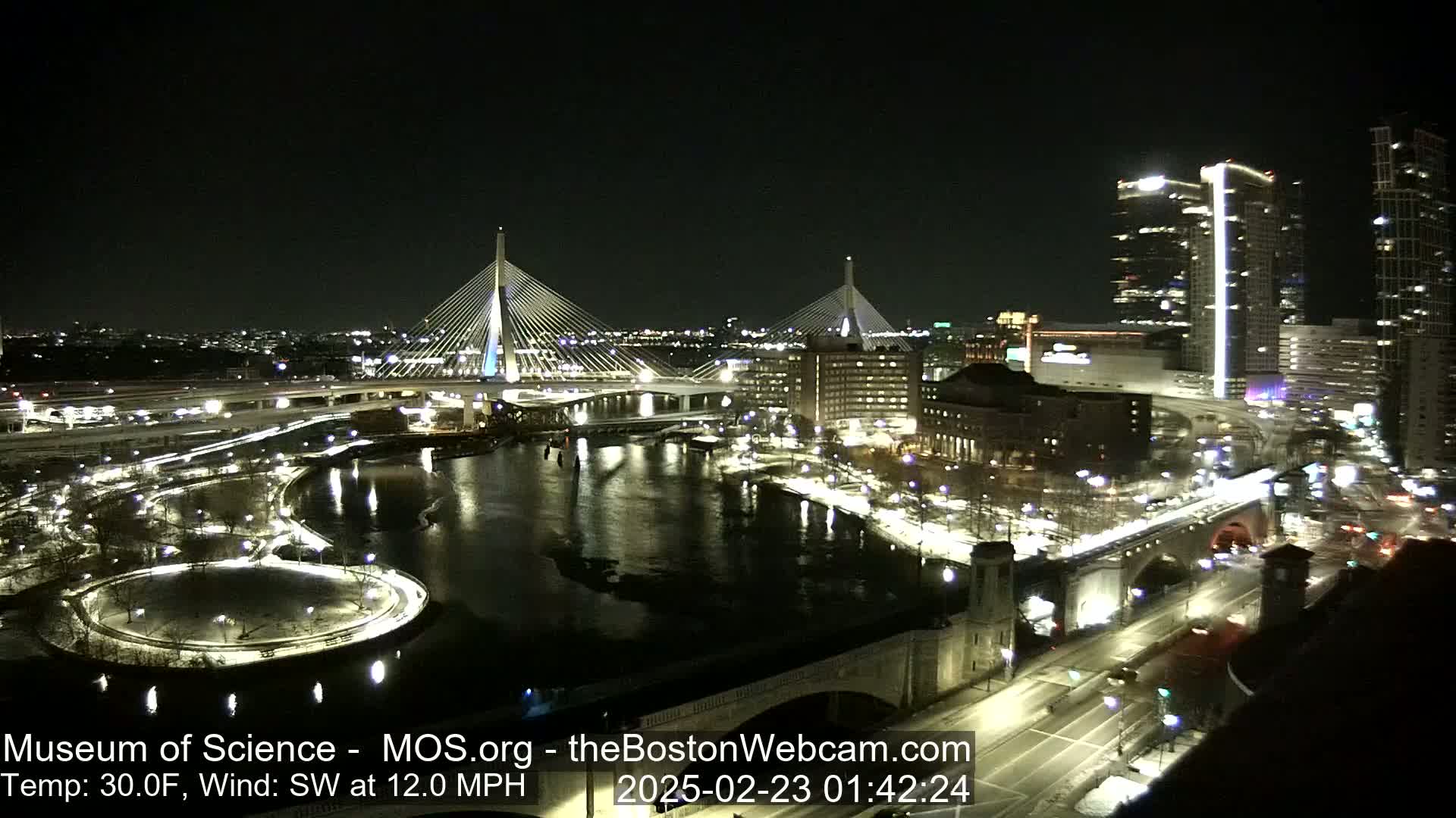 A nighttime cityscape with two cable-stayed bridges spanning a river, surrounded by city lights, roads, and snow-covered areas, under a clear sky with a temperature of 30°F and a southwest wind of 12 mph.