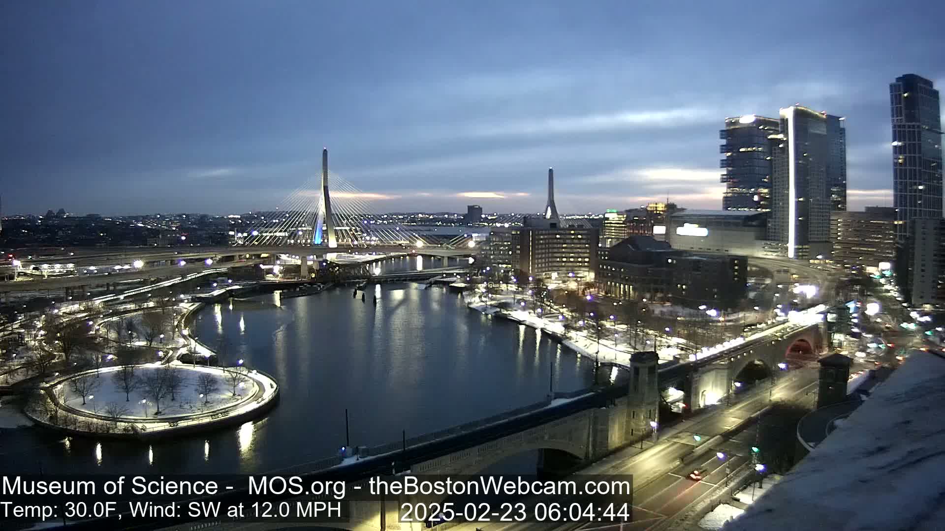 A nighttime cityscape with two cable-stayed bridges and a river, partially snow-covered, under a twilight sky with a southwest wind at 12 mph and a temperature of 30°F.