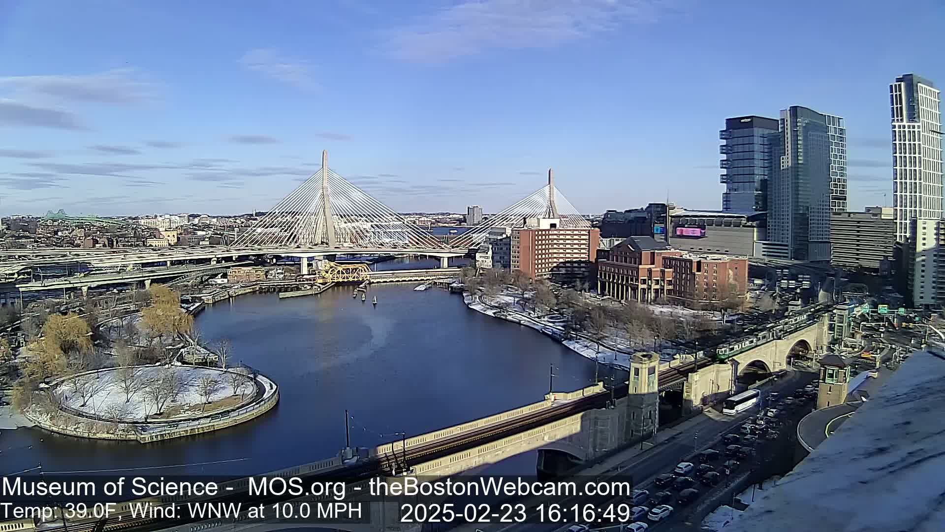 A sunny day with a light wind shows a city scene with two cable-stayed bridges spanning a river, alongside modern buildings and a train passing over a bridge, all lightly dusted with snow.