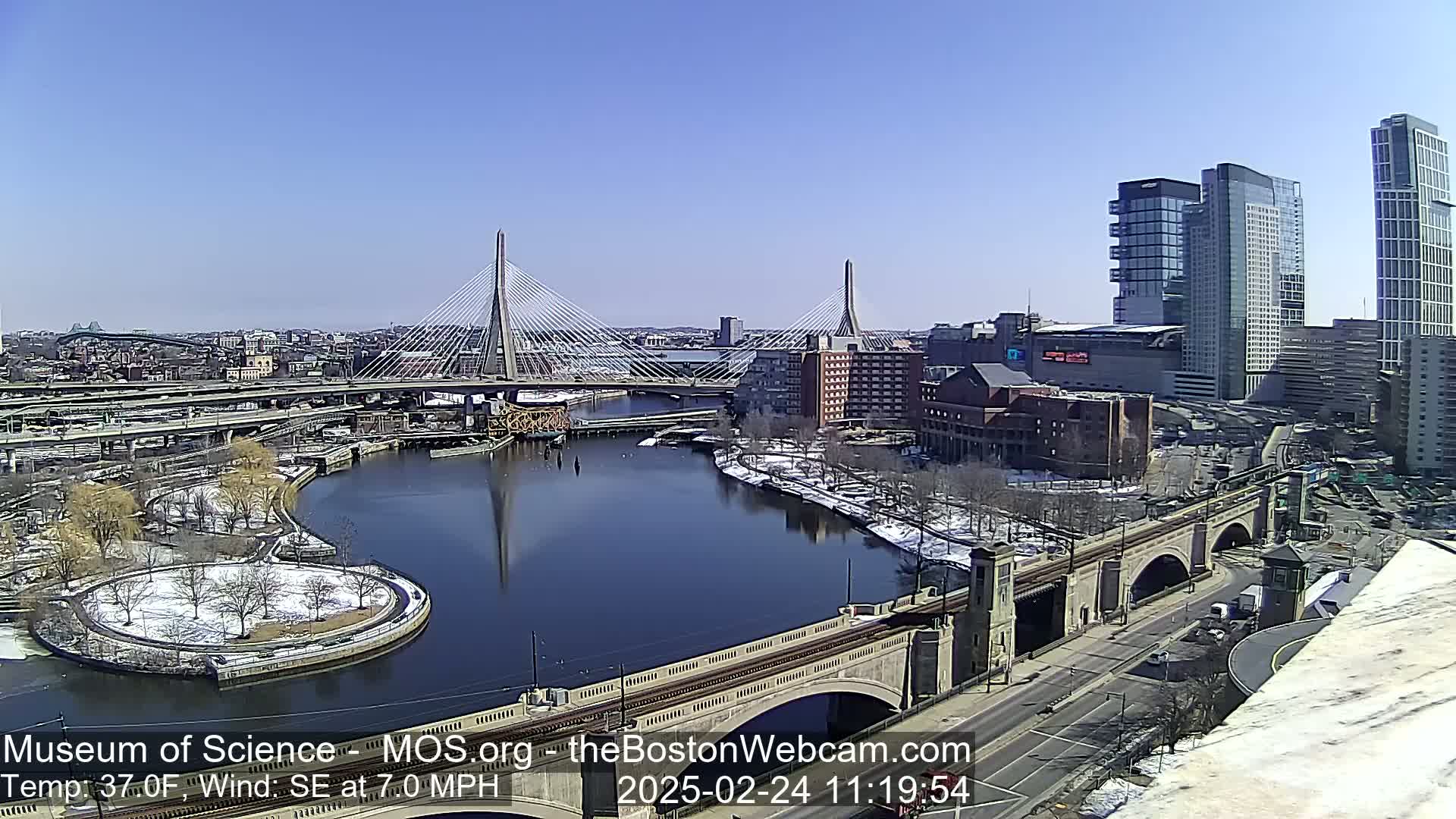 A sunny day reveals a cityscape with two cable-stayed bridges spanning a river, partially snow-covered land, and modern high-rise buildings.