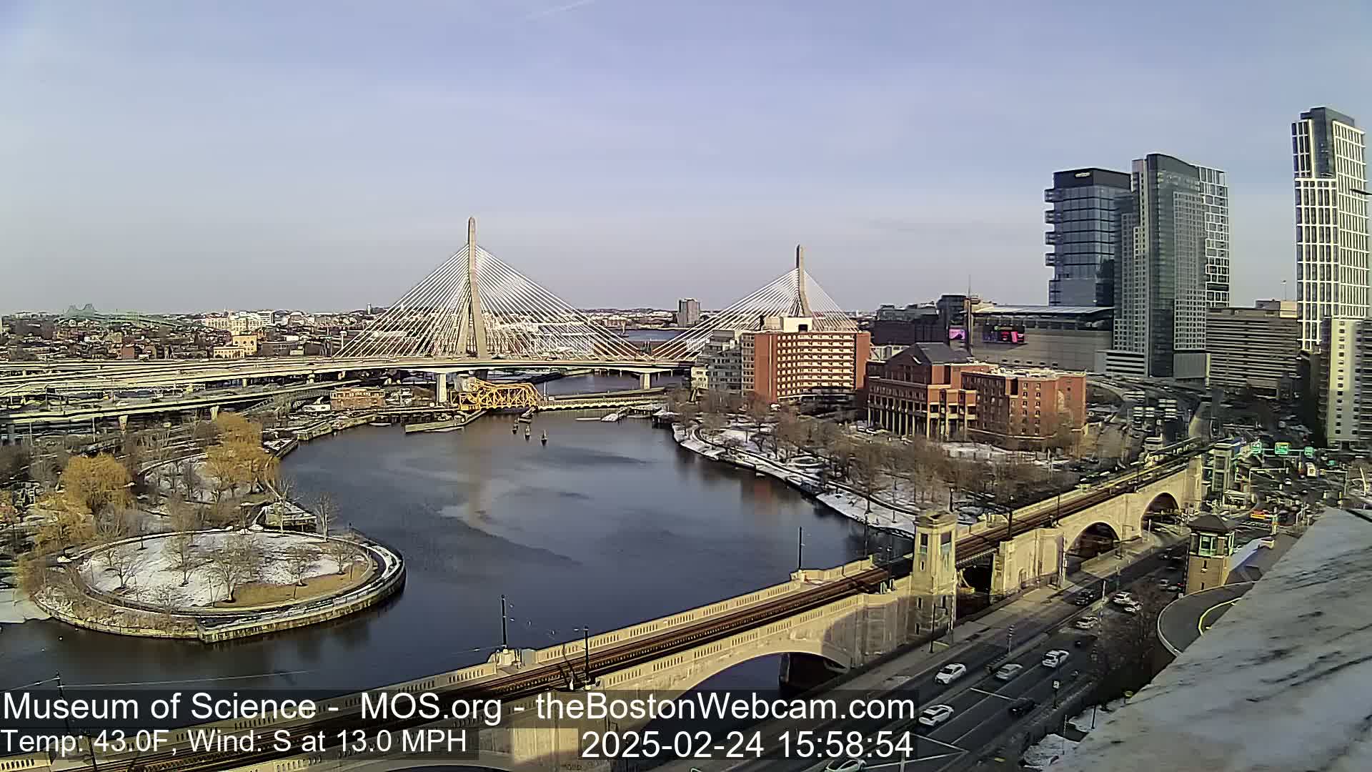 A sunny day with a 43°F temperature and a 13 mph south wind reveals a cityscape with two cable-stayed bridges spanning a river, alongside modern high-rise buildings and a park with patches of snow.