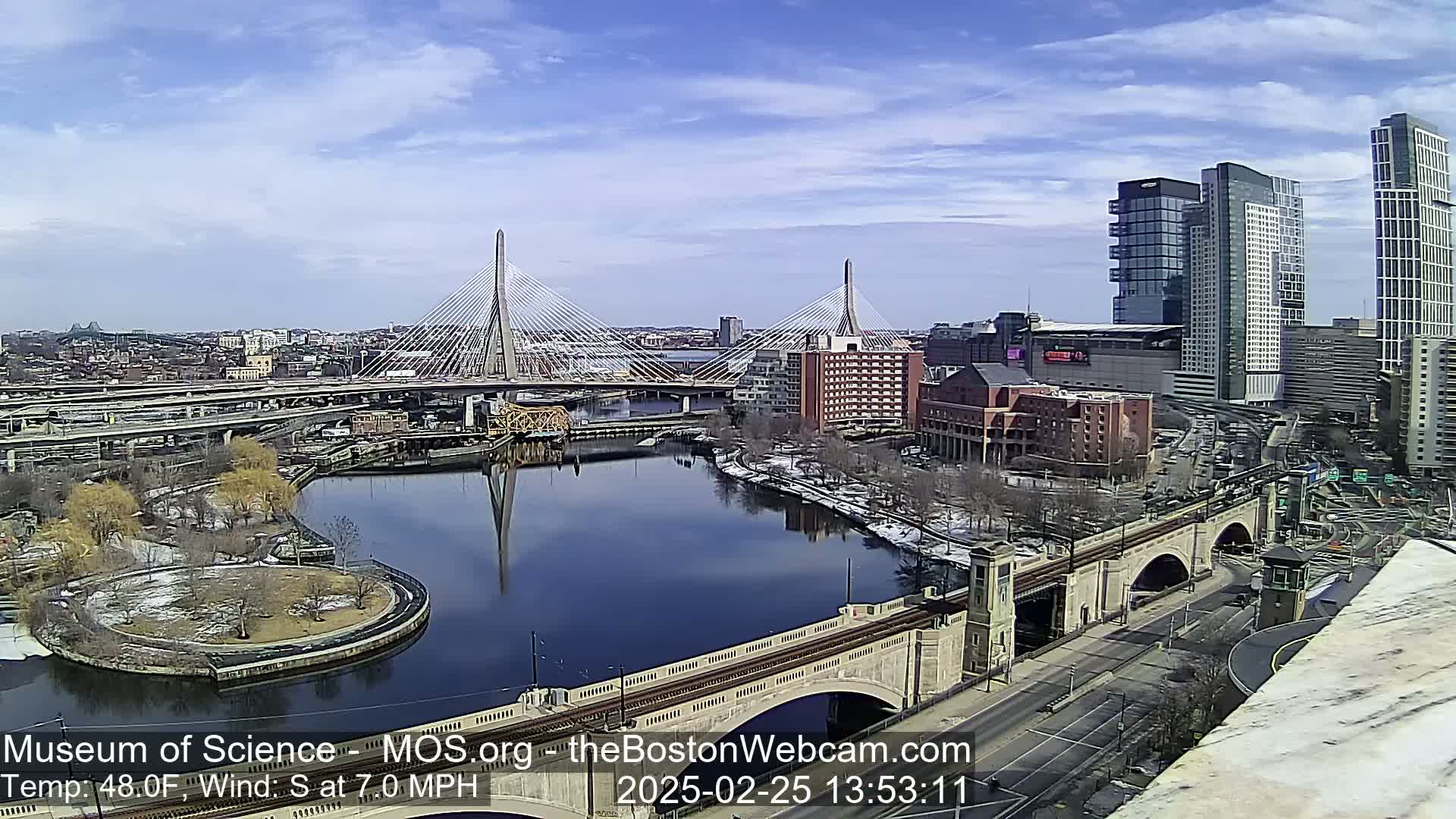 A sunny day with a temperature of 48°F and a south wind at 7 mph reveals a cityscape featuring two cable-stayed bridges spanning a river, with some remaining snow visible on the ground.