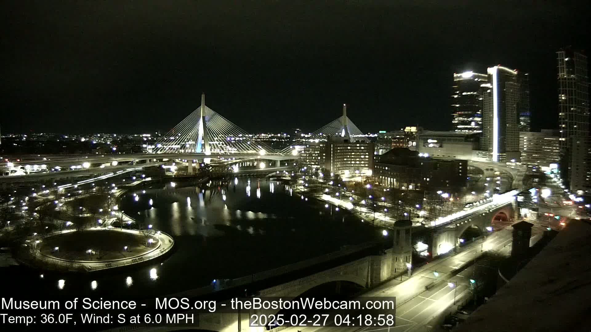 A nighttime, illuminated cityscape shows two cable-stayed bridges spanning a dark river, with city buildings and roadways visible under a clear sky and a temperature of 36 degrees Fahrenheit, with a 6 mph south wind.