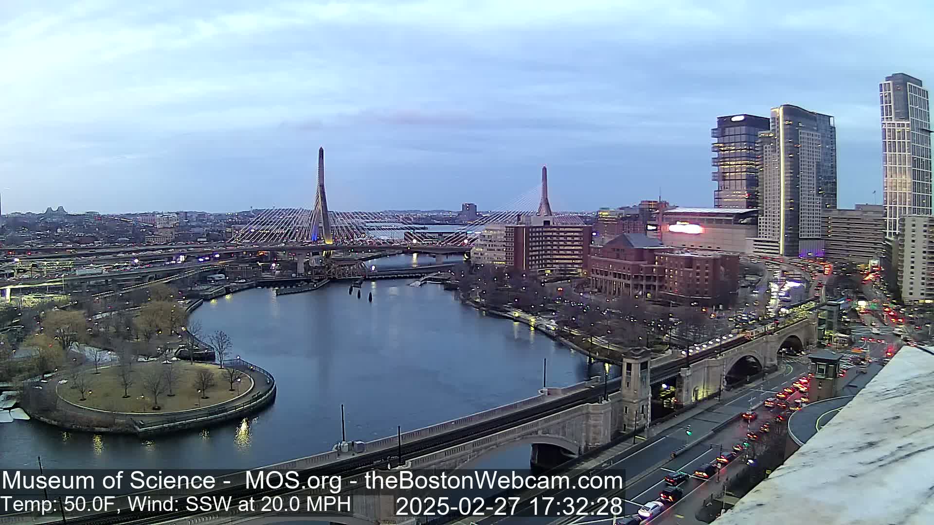 A twilight view of a city's waterfront, featuring two cable-stayed bridges, a river, and several buildings, under a mostly clear sky with a 20 mph south-southwesterly wind and a temperature of 50 degrees Fahrenheit.