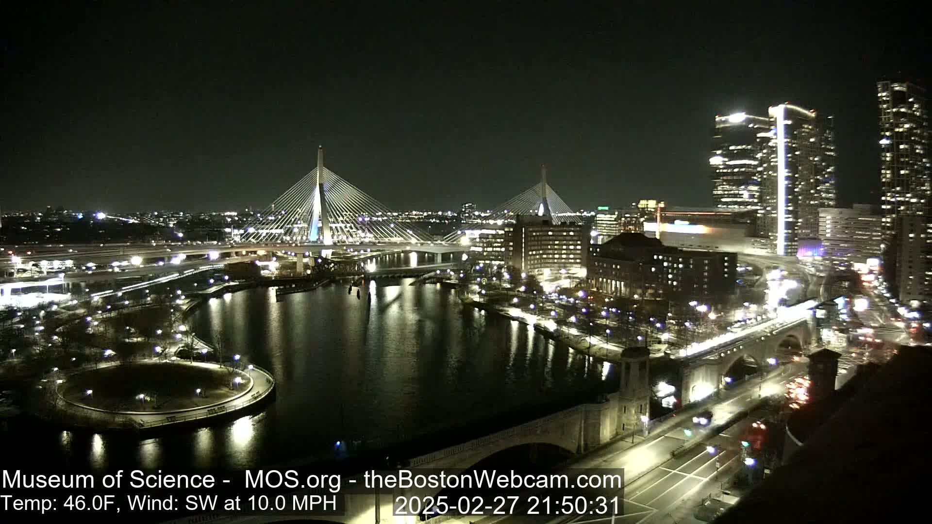 A nighttime cityscape showing two cable-stayed bridges spanning a dark river, with city lights reflecting on the water under a clear sky, and a southwest wind of 10 mph.