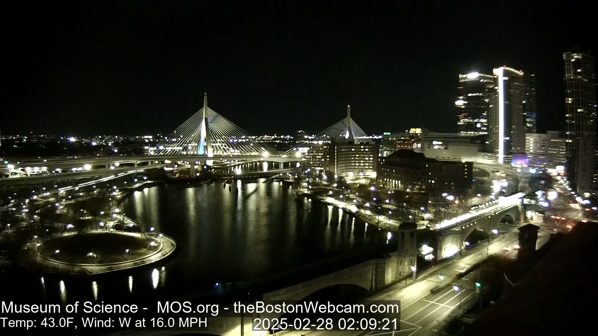 A nighttime cityscape with two cable-stayed bridges spanning a dark river, under a clear sky with a 43°F temperature and a 16 mph west wind.