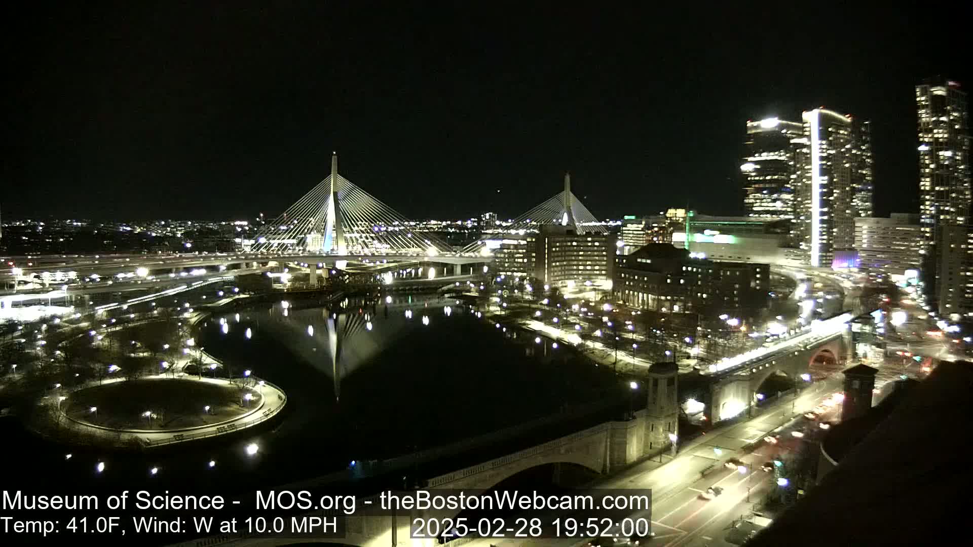 A nighttime view of two illuminated cable-stayed bridges spanning a dark river, surrounded by city lights and buildings under a clear sky.