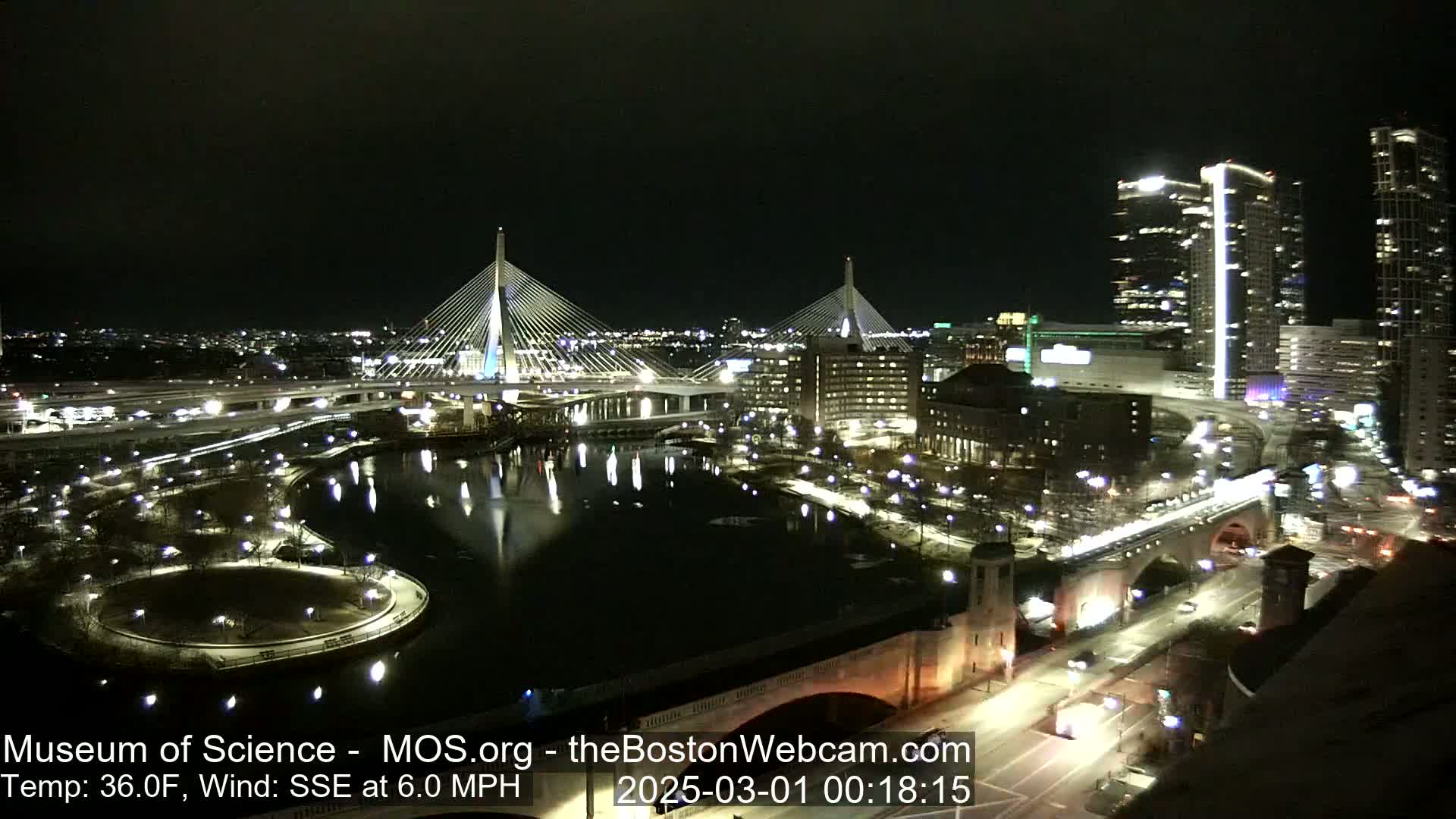 A nighttime cityscape with two illuminated cable-stayed bridges spanning a dark river, surrounded by city lights and buildings, under a clear sky with a temperature of 36°F and a southeasterly wind of 6.0 mph.