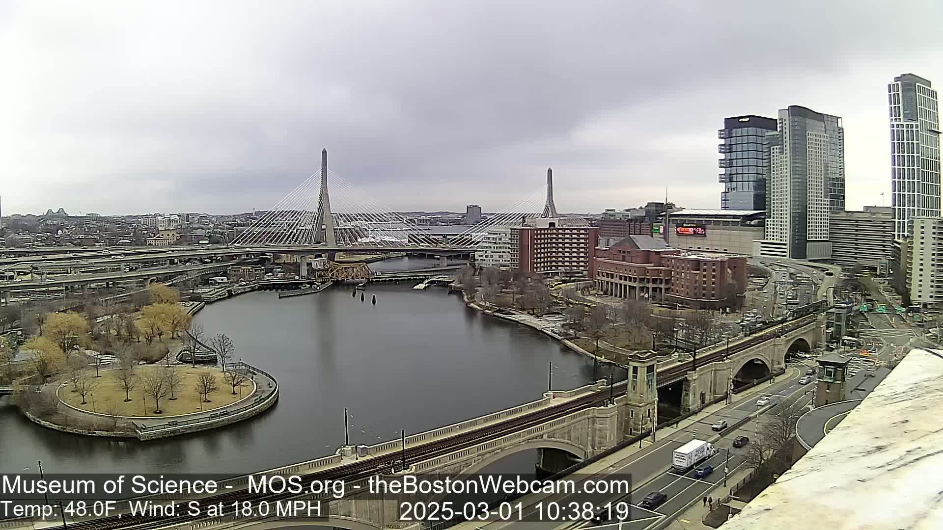 A city river scene under an overcast sky shows two cable-stayed bridges, a park, and modern high-rise buildings.