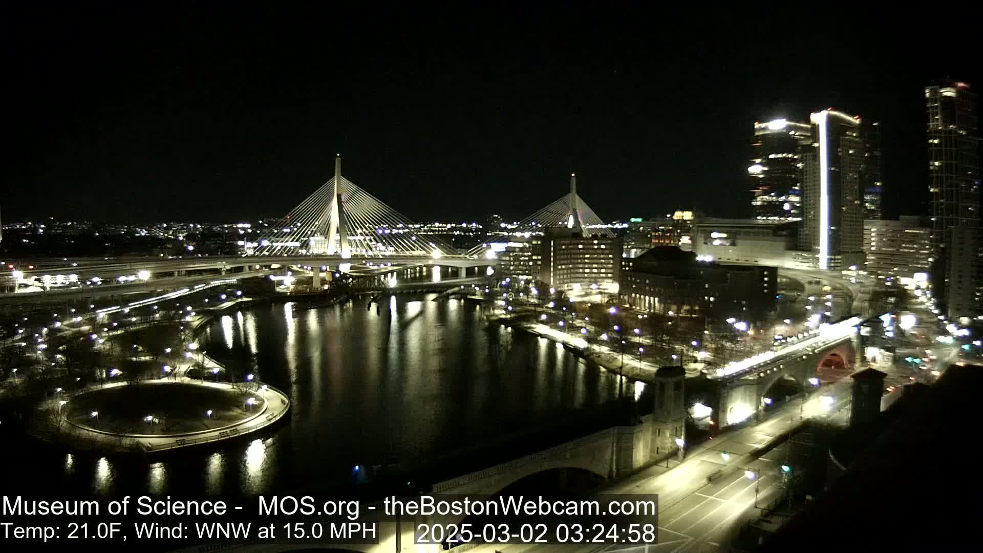 A nighttime aerial view of a city with two illuminated cable-stayed bridges spanning a dark river, under a clear sky with a temperature of 21°F and a wind of 15 mph from the west-northwest.