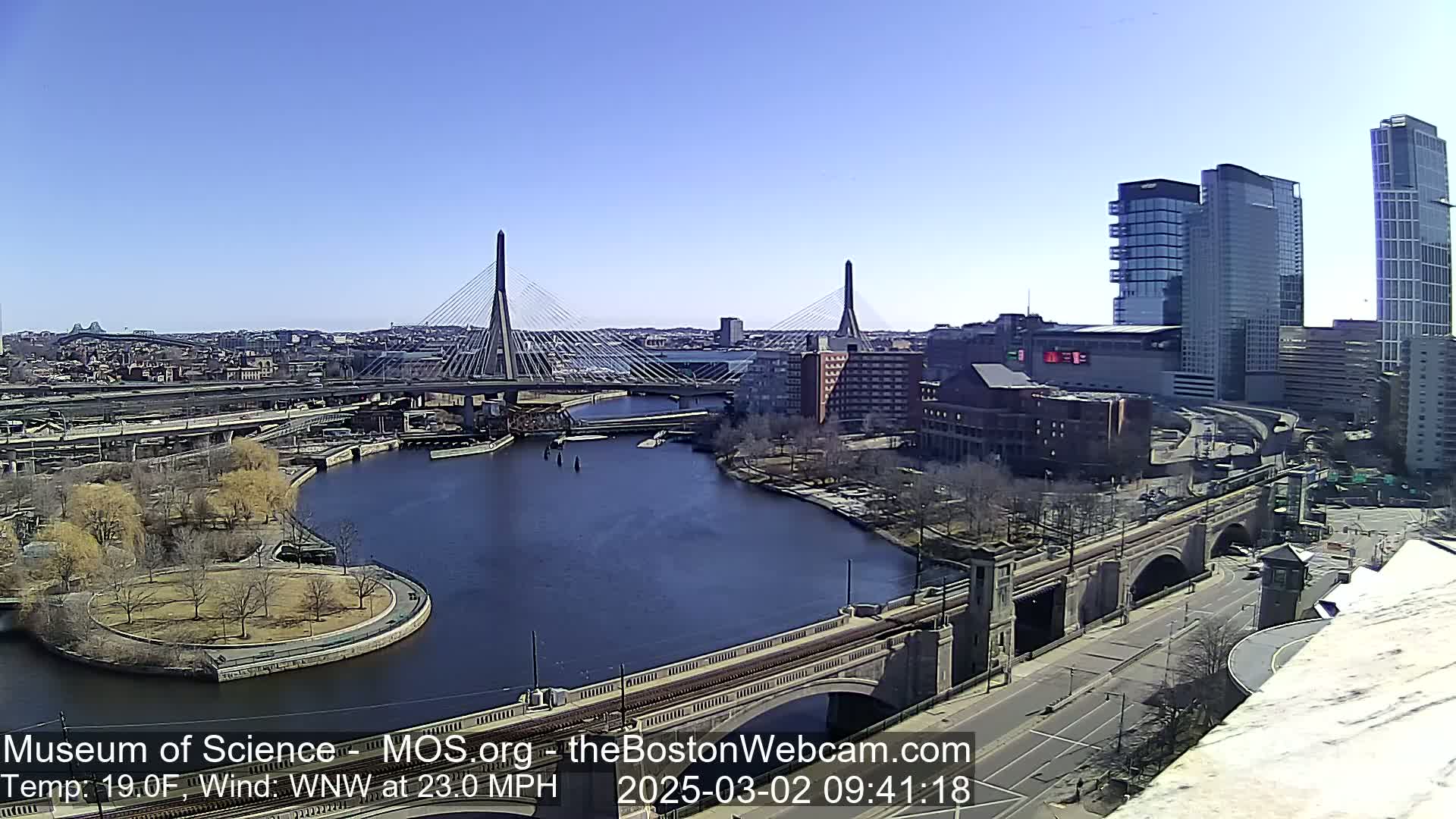 A sunny day with a 23-mph wind reveals a cityscape with two cable-stayed bridges spanning a river, alongside other bridges and modern high-rise buildings.