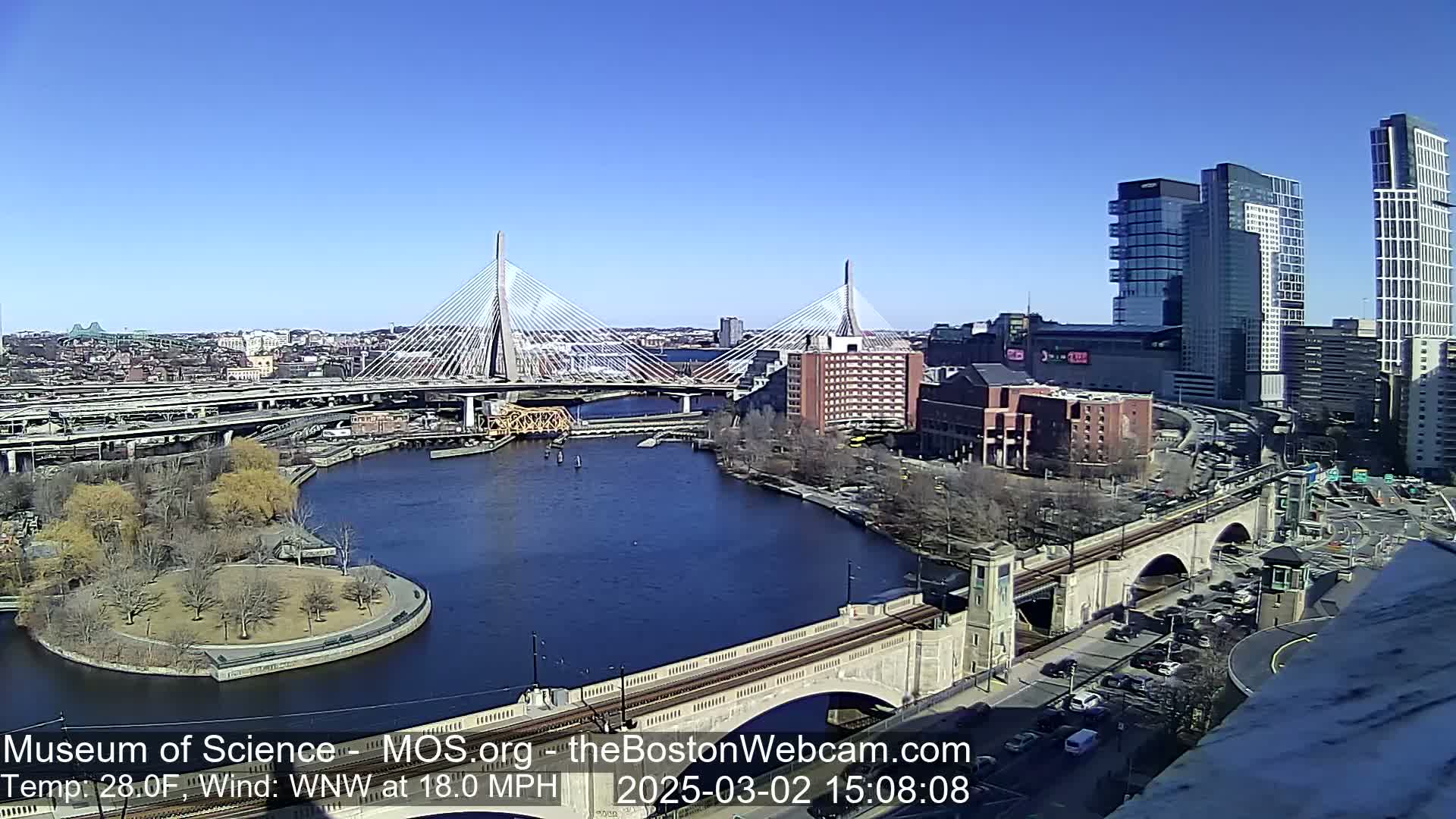 A sunny cityscape with two cable-stayed bridges spanning a river, surrounded by buildings, roadways, and a park, under a clear blue sky with a wind of 18 mph from the west-northwest.