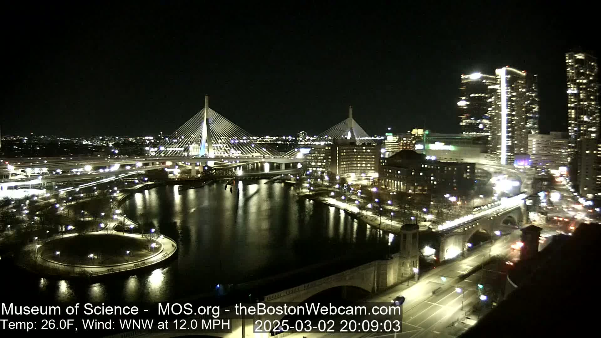 A nighttime cityscape featuring two illuminated cable-stayed bridges spanning a dark river, with city buildings and streets also lit, under a clear sky and a 26°F temperature with a 12 mph wind.