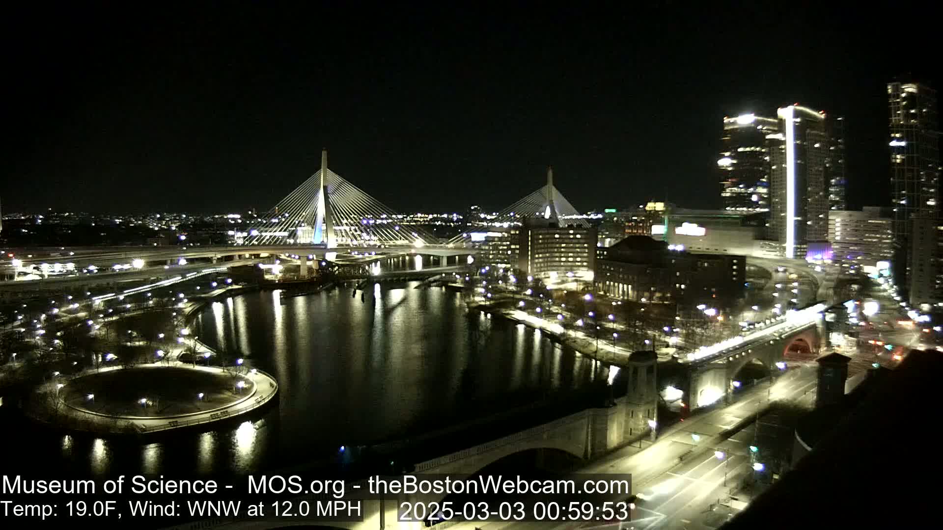 A nighttime view of two illuminated cable-stayed bridges over a dark river, surrounded by city lights and buildings, under a clear sky with a 19°F temperature and a 12 mph wind from the west-northwest.