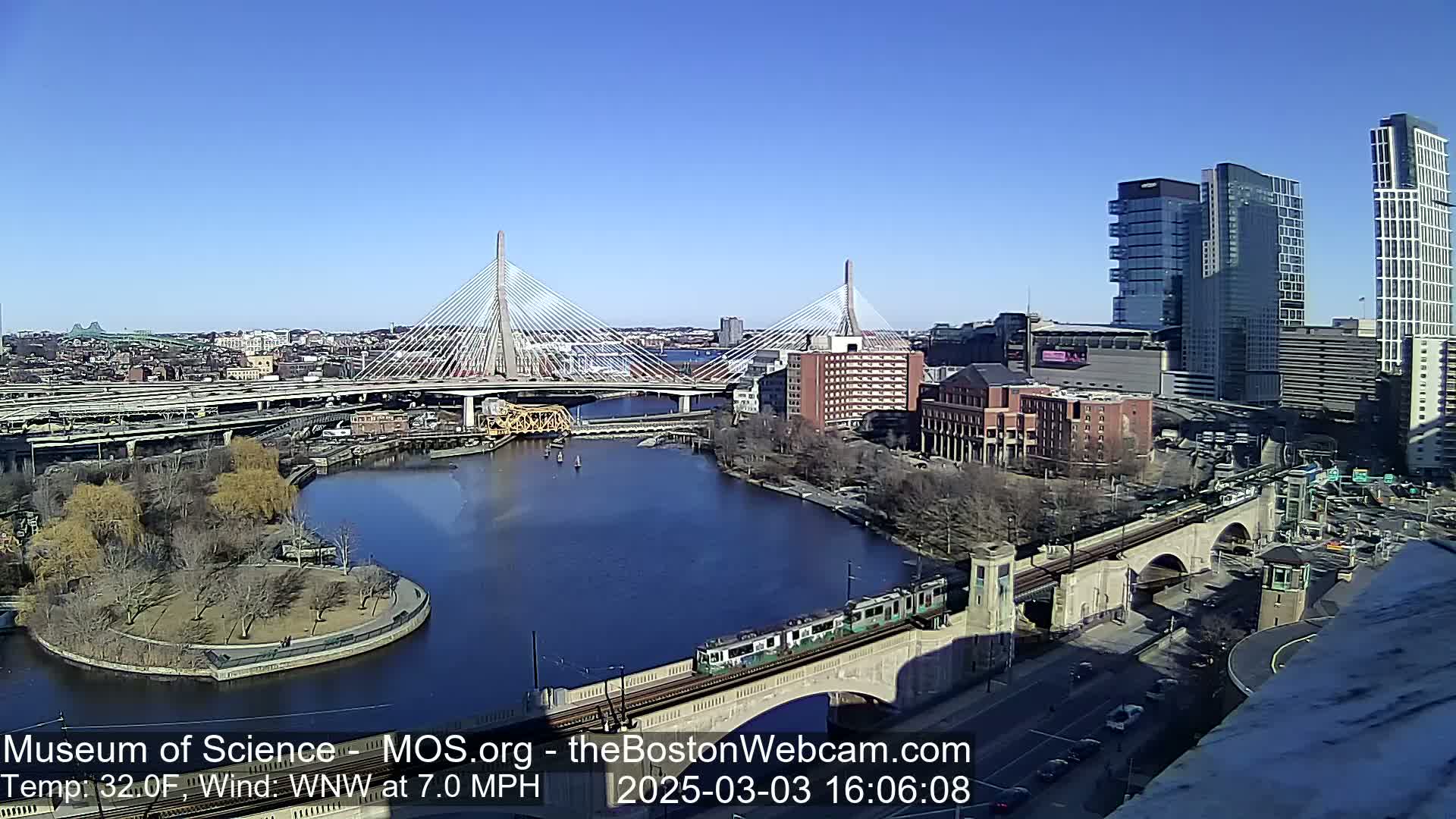 A sunny day shows a city with a river, two cable-stayed bridges, and a train crossing a stone bridge.