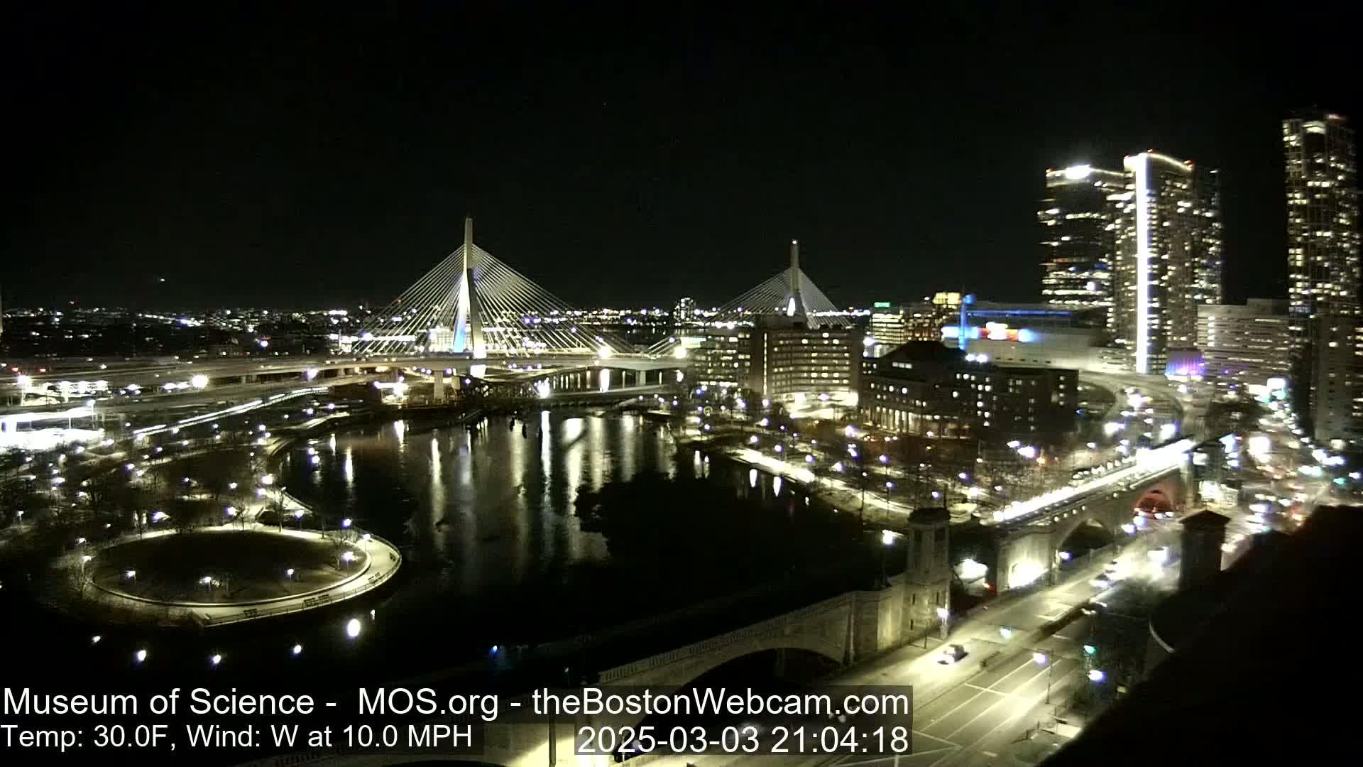 A nighttime view of a city with two illuminated cable-stayed bridges spanning a dark river, surrounded by buildings and roads with traffic under a clear sky, with a temperature of 30°F and a 10 mph westerly wind.