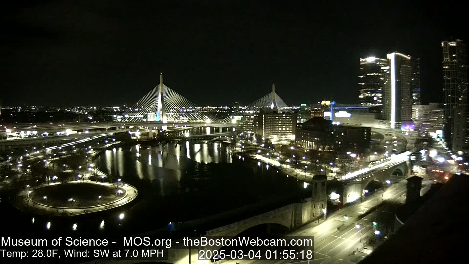 A nighttime cityscape featuring two illuminated cable-stayed bridges spanning a dark river, with city lights and buildings reflecting in the water, under a clear night sky with a southwest wind at 7 mph and a temperature of 28 degrees Fahrenheit.