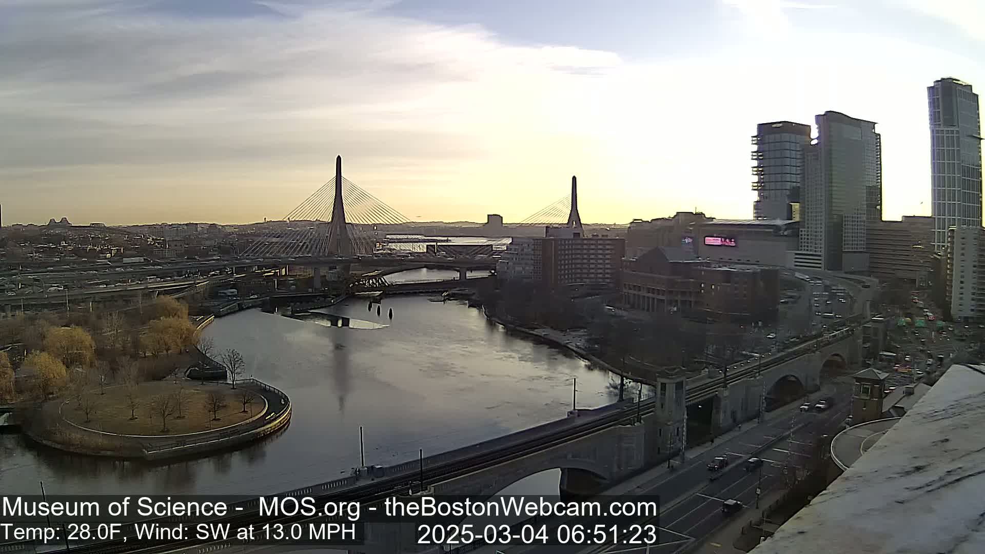 A cityscape at sunrise with two cable-stayed bridges spanning a river, under partly cloudy skies and a southwest wind of 13 mph.