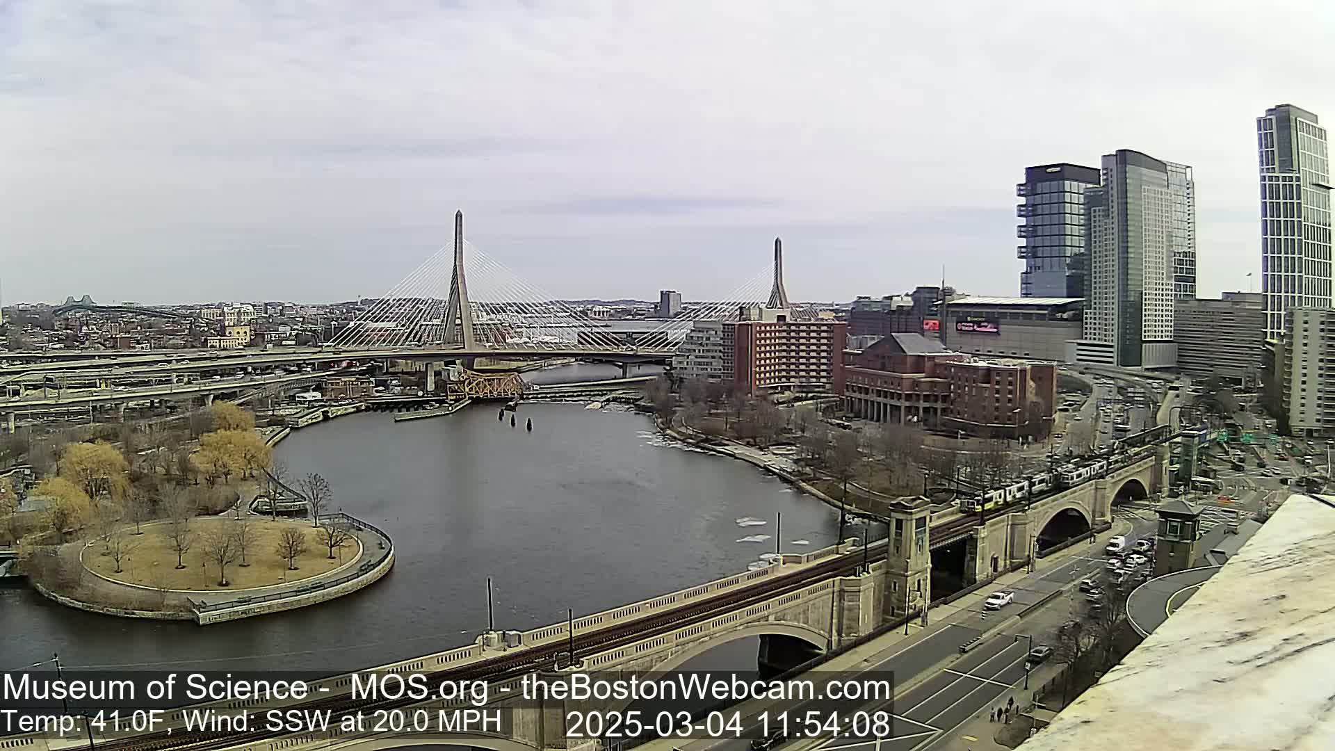 A city scene on a partly cloudy day with a 20 mph wind shows two cable-stayed bridges spanning a river, with modern high-rise buildings and a train visible in the background.