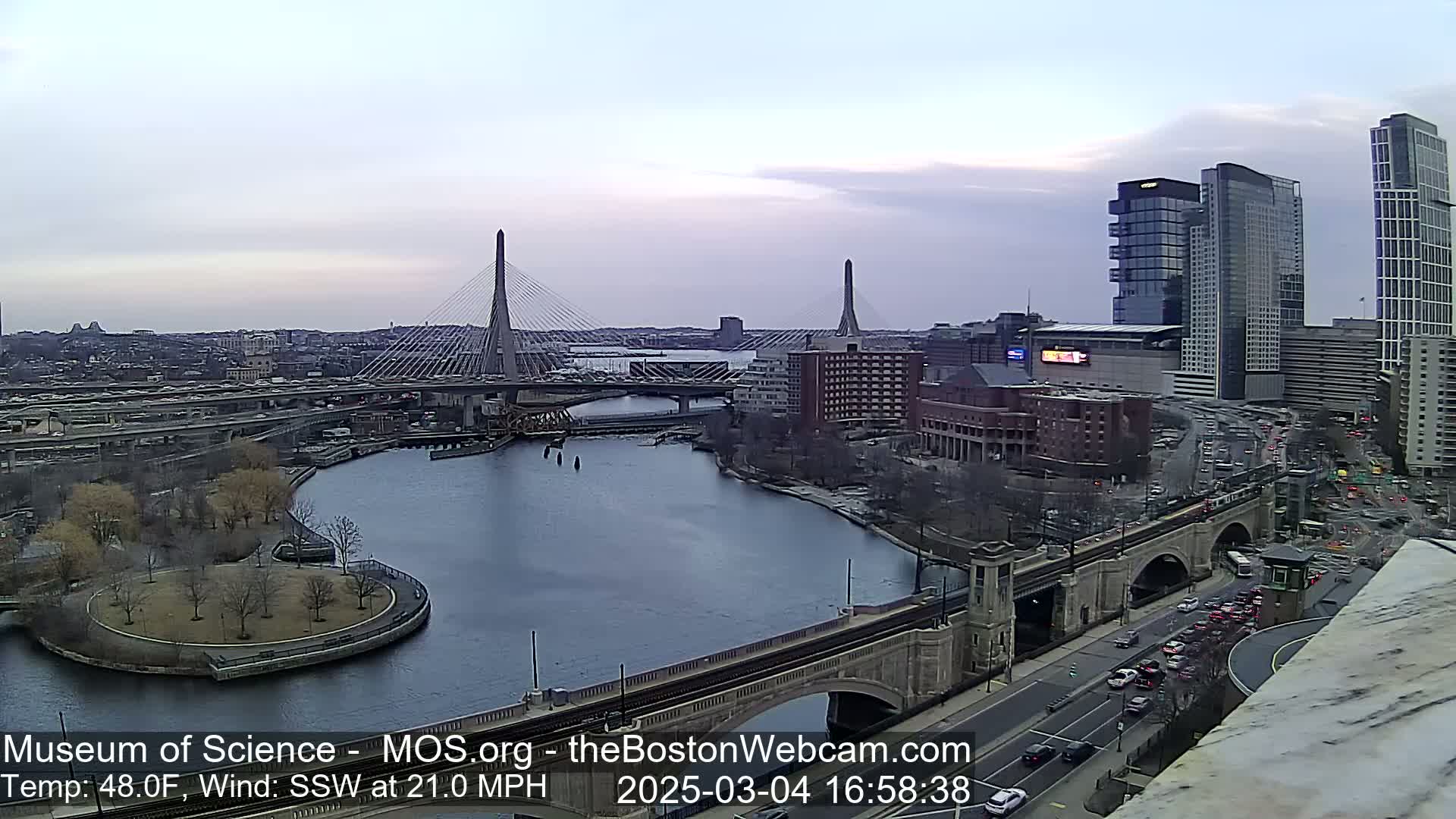 A cityscape with two cable-stayed bridges over a river, under a partly cloudy sky, with a 48°F temperature and a 21 mph southwest wind.