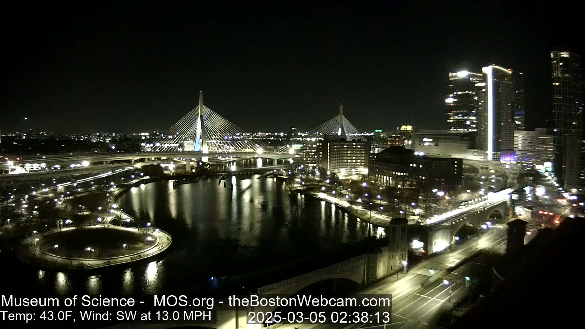 A nighttime cityscape featuring two illuminated cable-stayed bridges spanning a dark river, under a clear sky with a temperature of 43°F and a southwest wind of 13 mph.