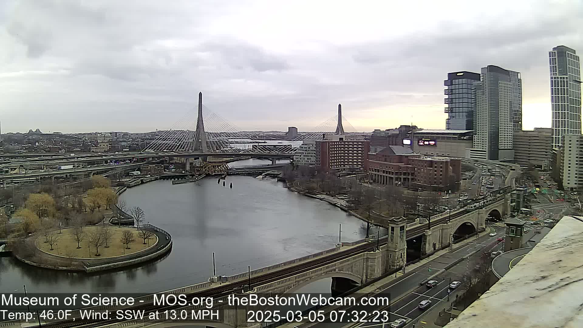 A city river scene under an overcast sky shows two cable-stayed bridges, a park, and several buildings, including high-rises.