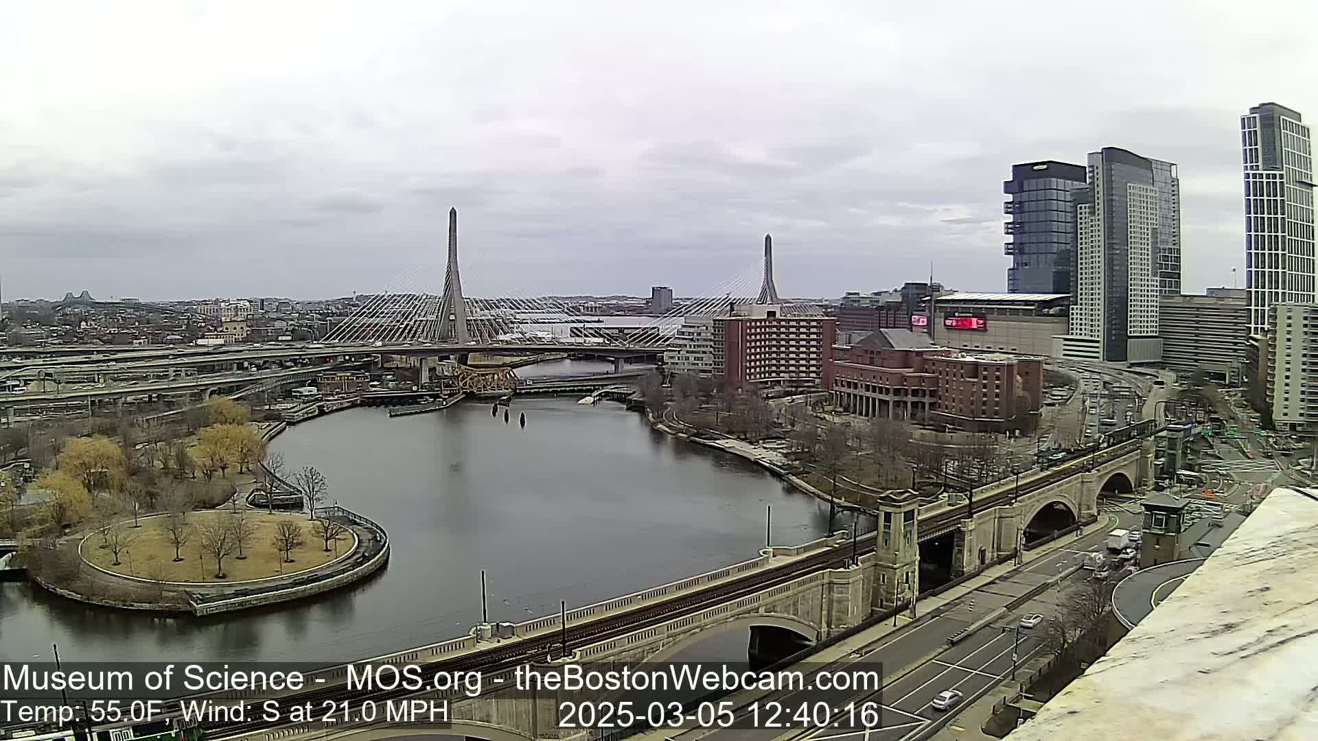 A city landscape with two cable-stayed bridges over a river, under an overcast sky.