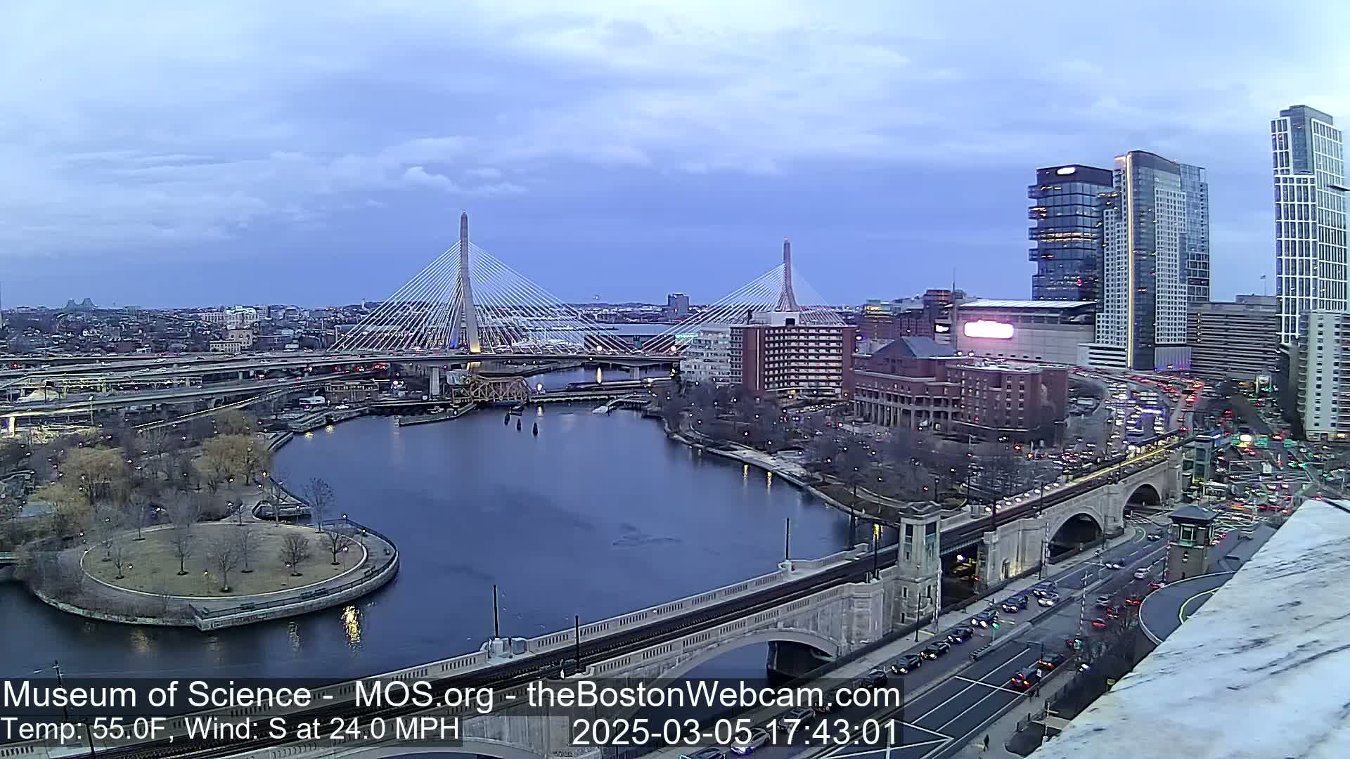 A view of a city at dusk showing two cable-stayed bridges spanning a river, with a city park and several buildings, including tall skyscrapers, under a mostly cloudy sky with a south wind at 24 mph.