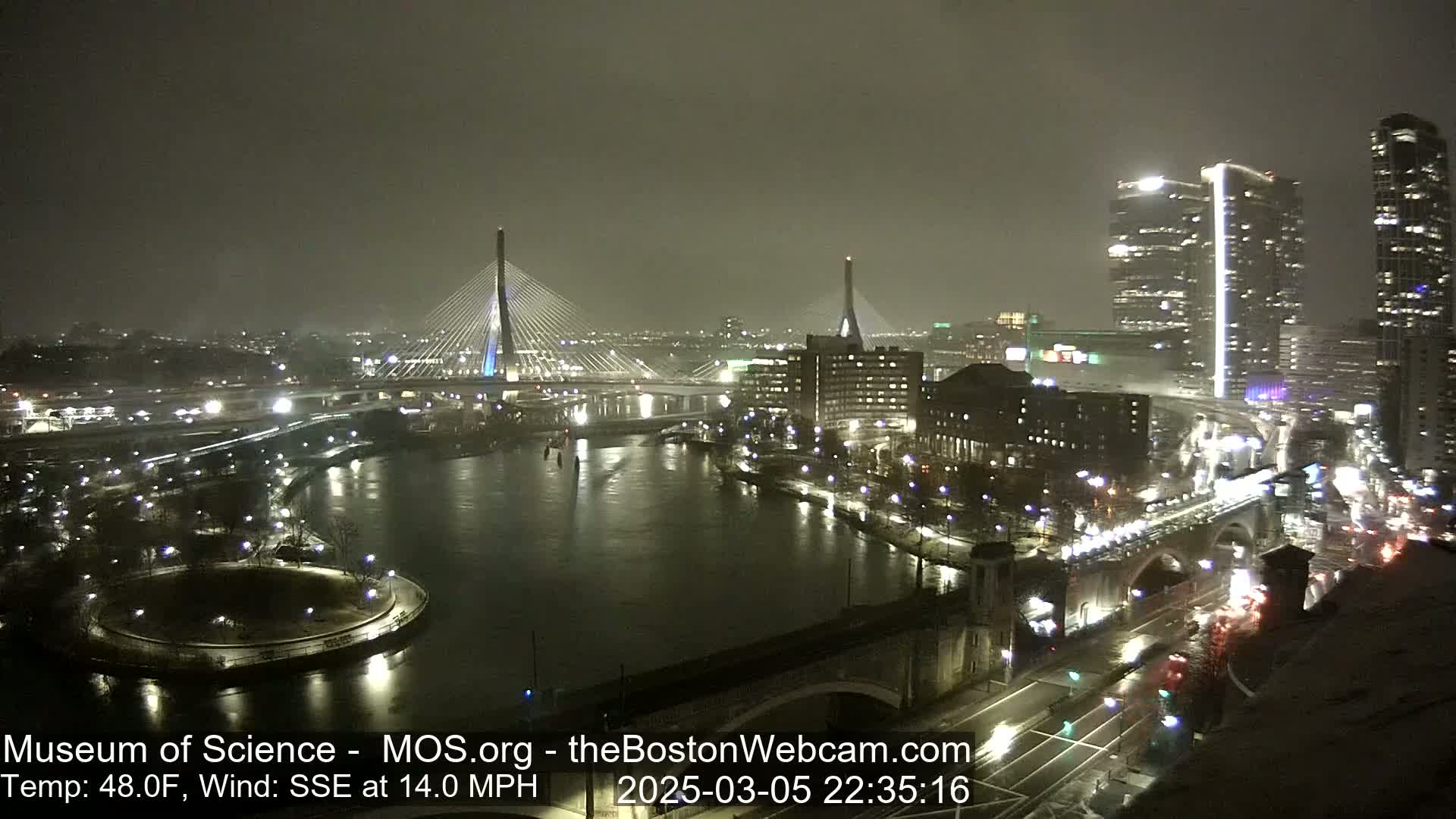 A nighttime view of a city illuminated with lights, featuring two cable-stayed bridges spanning a river, under a dark, possibly snowy or rainy sky.