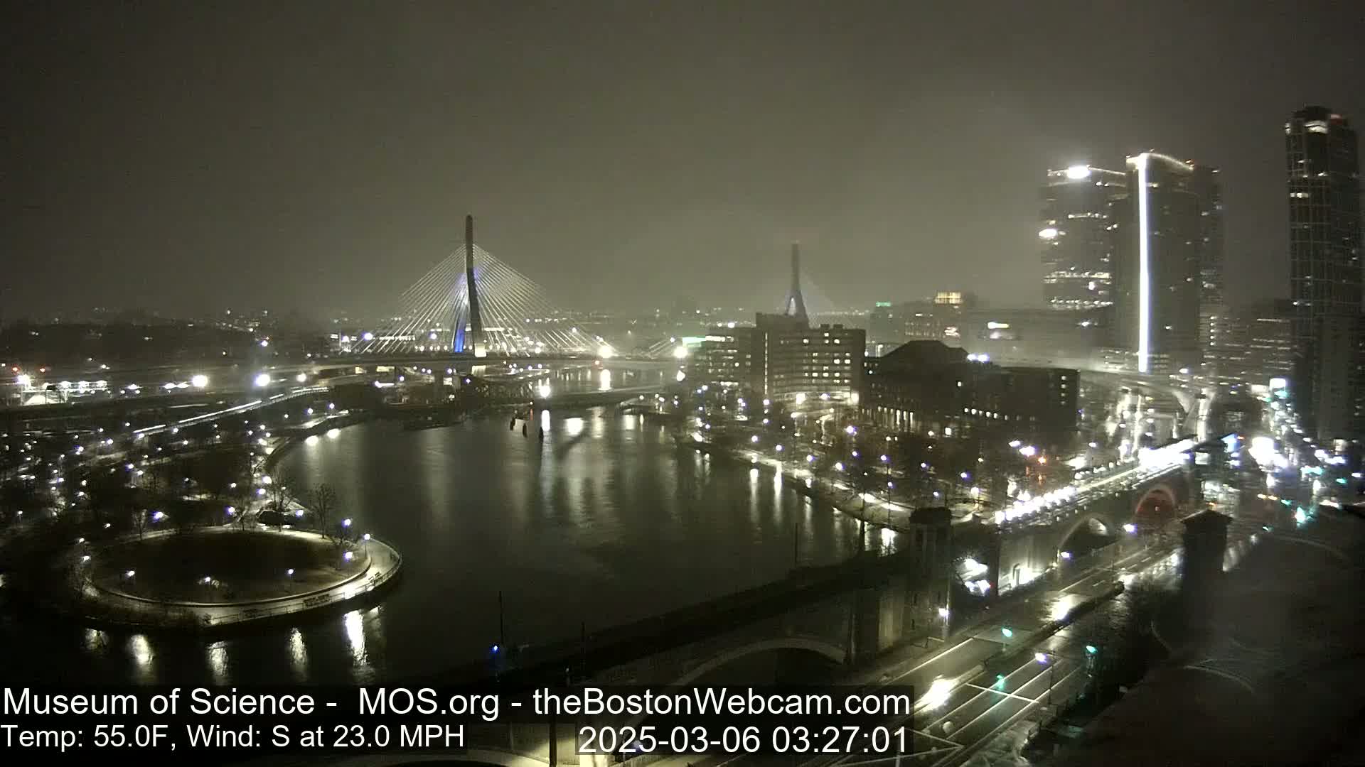 A nighttime view of a city illuminated by lights, featuring two cable-stayed bridges spanning a river under hazy, windy conditions.