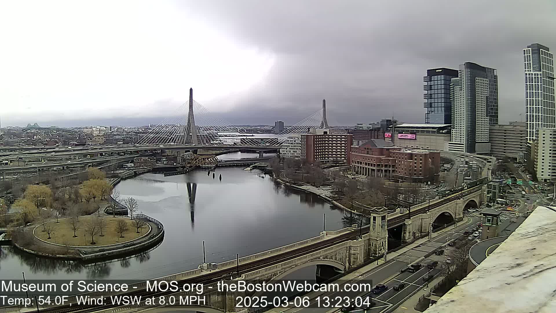 A city river scene on an overcast day shows two cable-stayed bridges, a roadway, and modern high-rise buildings.