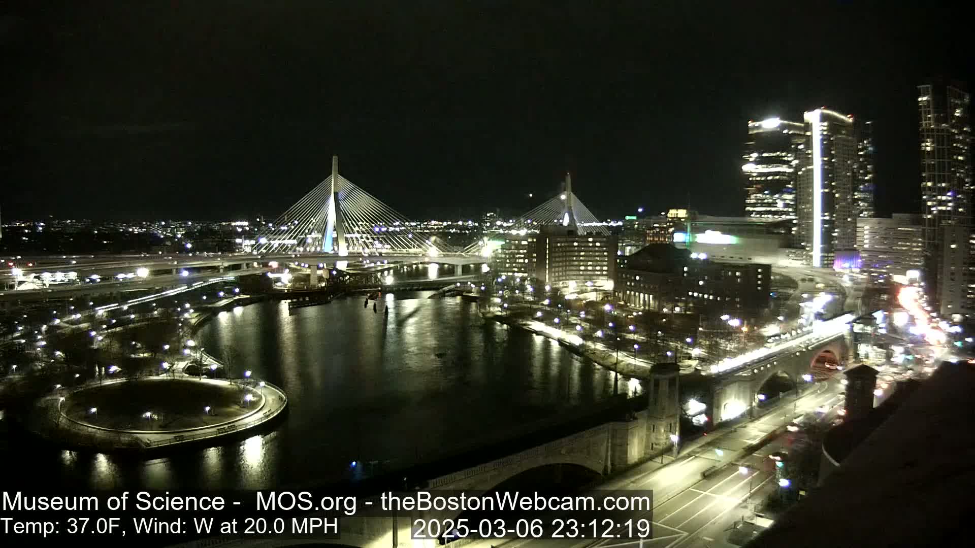 A nighttime view of two cable-stayed bridges spanning a river, with city lights illuminating buildings and roads, under a clear, windy sky with a temperature of 37 degrees Fahrenheit.