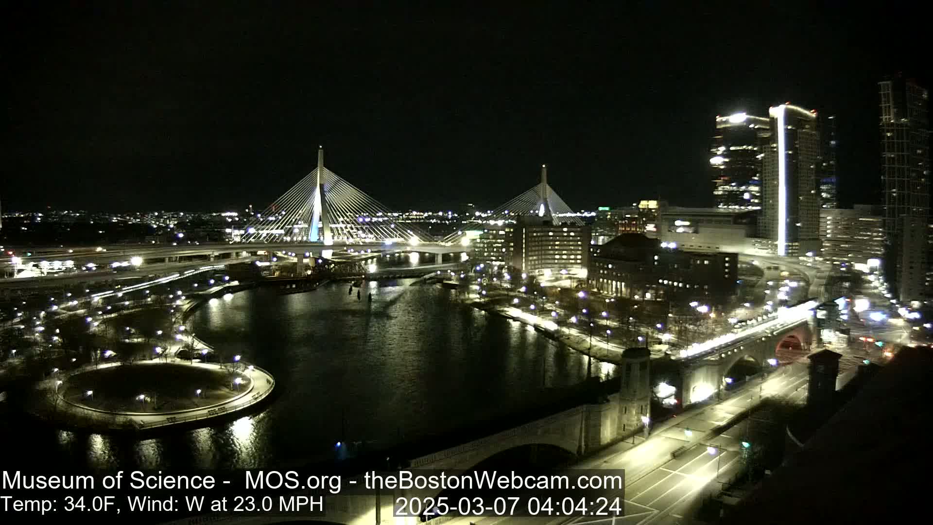 A nighttime cityscape with two illuminated cable-stayed bridges spanning a dark river, under a clear sky and a 23 mph west wind.