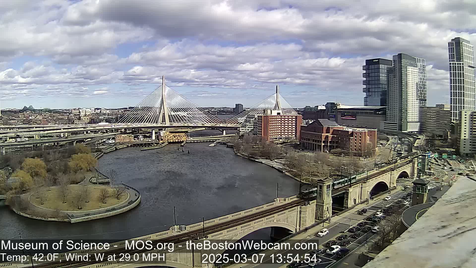 A partly cloudy day with a westerly wind of 29 mph shows two cable-stayed bridges spanning a river, with city buildings and a train track in the foreground.