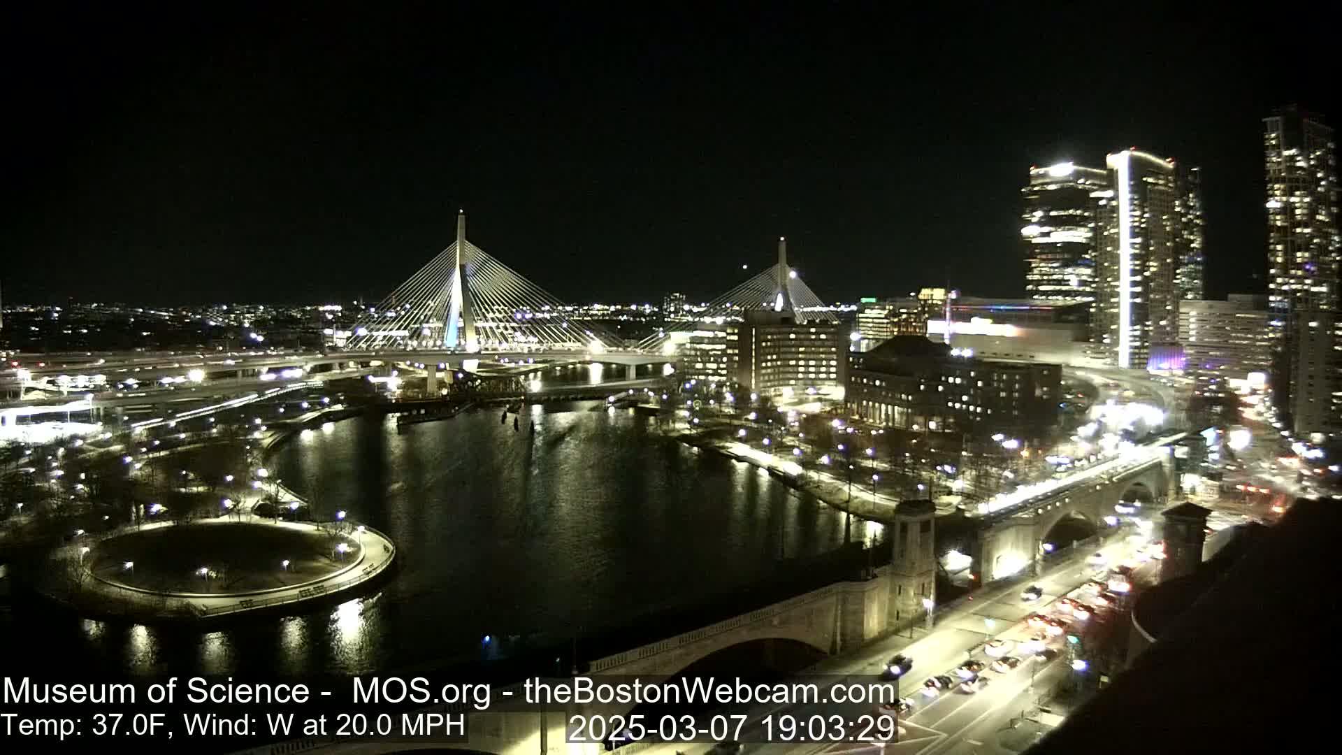 A nighttime aerial view of a city with two cable-stayed bridges spanning a dark river, under a clear sky with a 37°F temperature and 20 mph westerly wind.