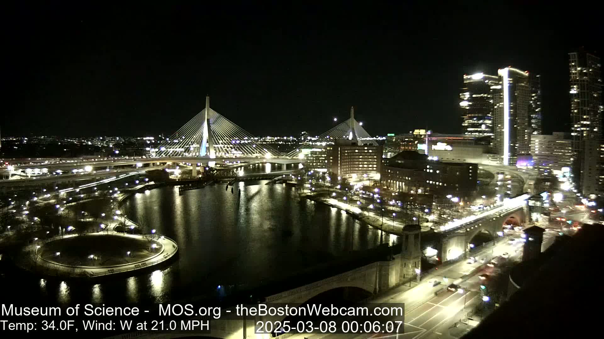 A nighttime view of a city waterfront, showing two illuminated cable-stayed bridges, a river, and city buildings, with a 34°F temperature and a 21 mph west wind.