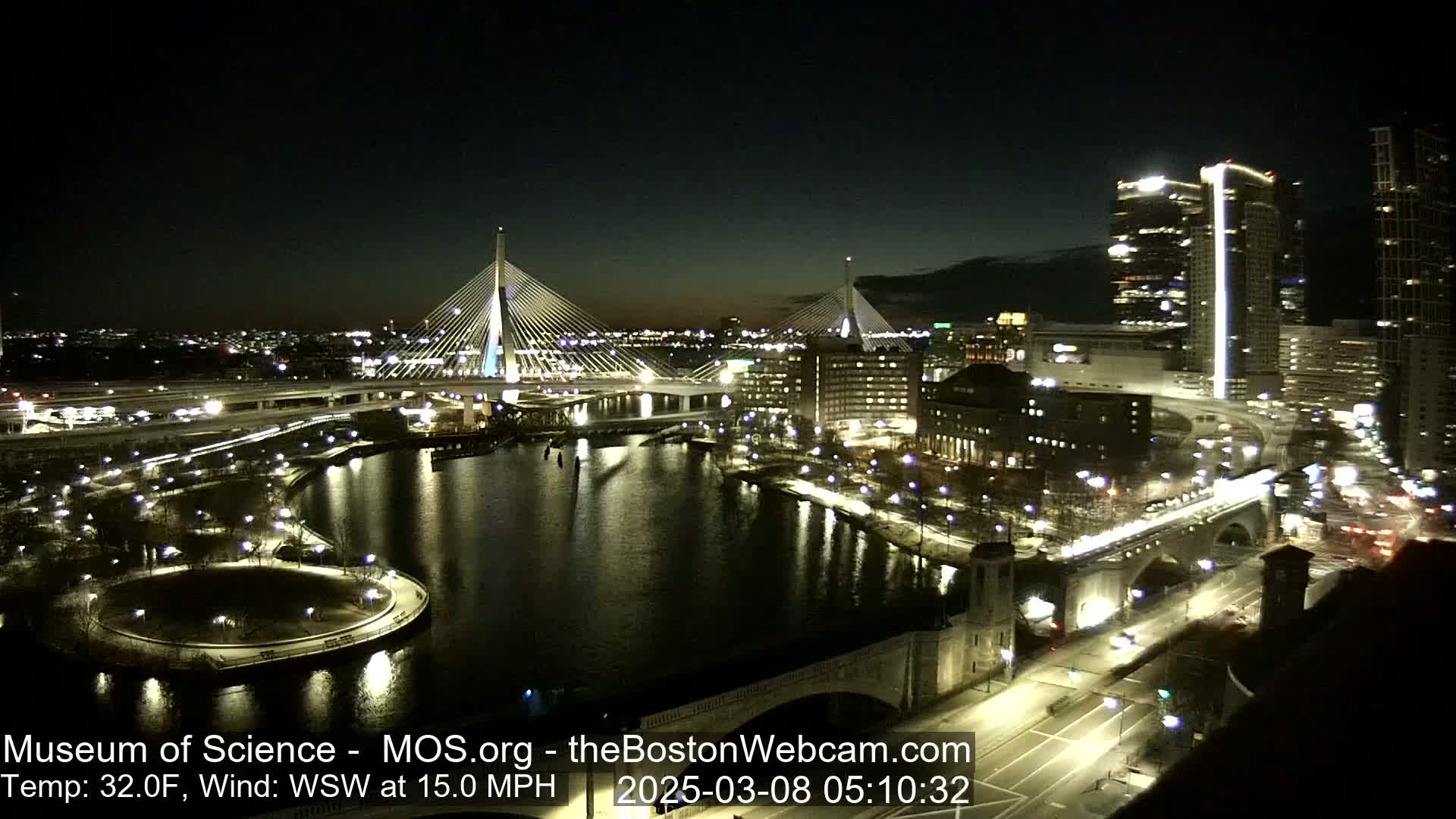 A nighttime, aerial view of a city with two illuminated cable-stayed bridges spanning a dark river, under a clear sky with a 32°F temperature and a 15 mph wind from the west-southwest.