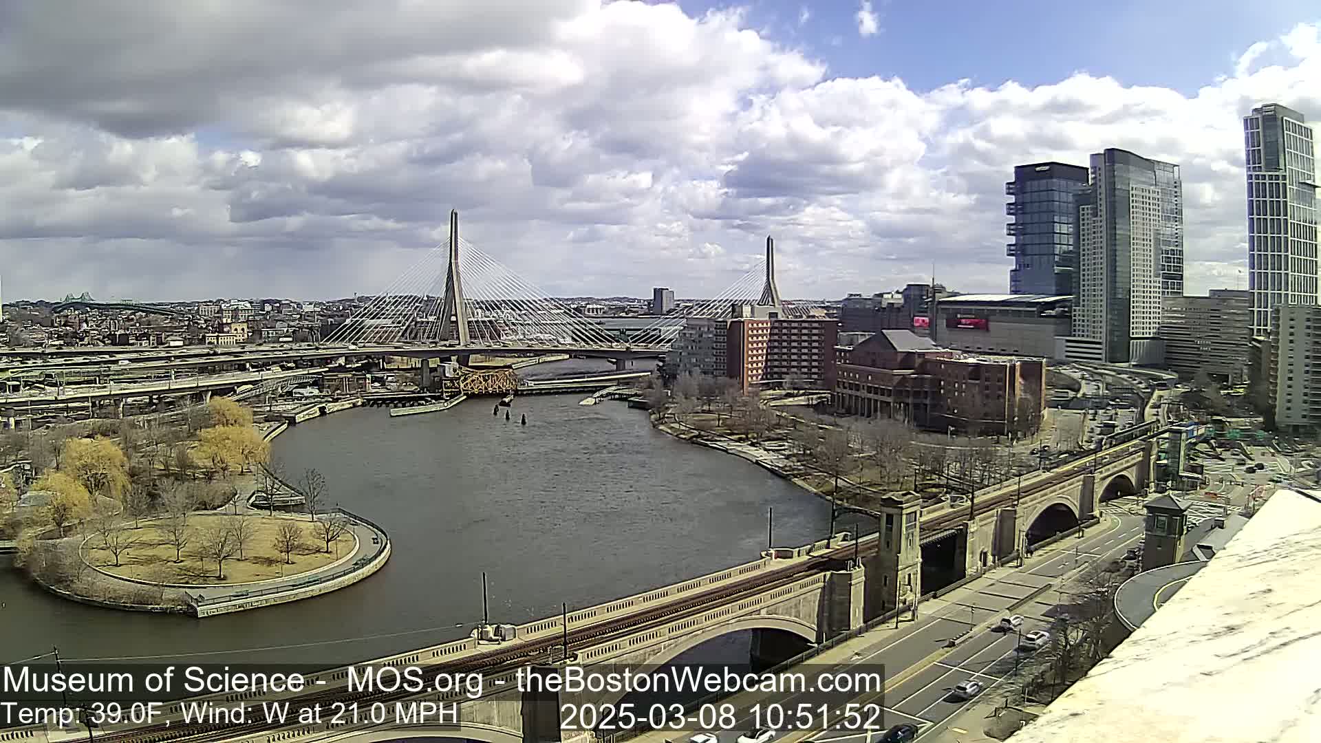 A partly cloudy day shows a cityscape with two cable-stayed bridges crossing a river, alongside other bridges and high-rise buildings.