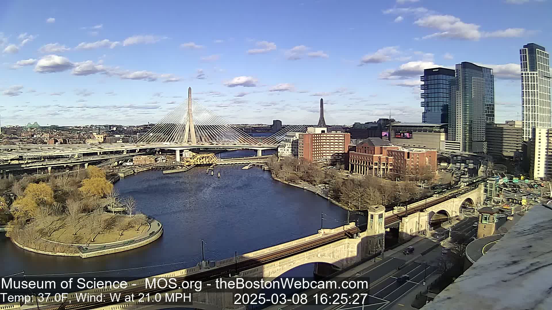 A sunny day with a westerly wind of 21 mph shows a cityscape with a cable-stayed bridge spanning a river, alongside other bridges and high-rise buildings.