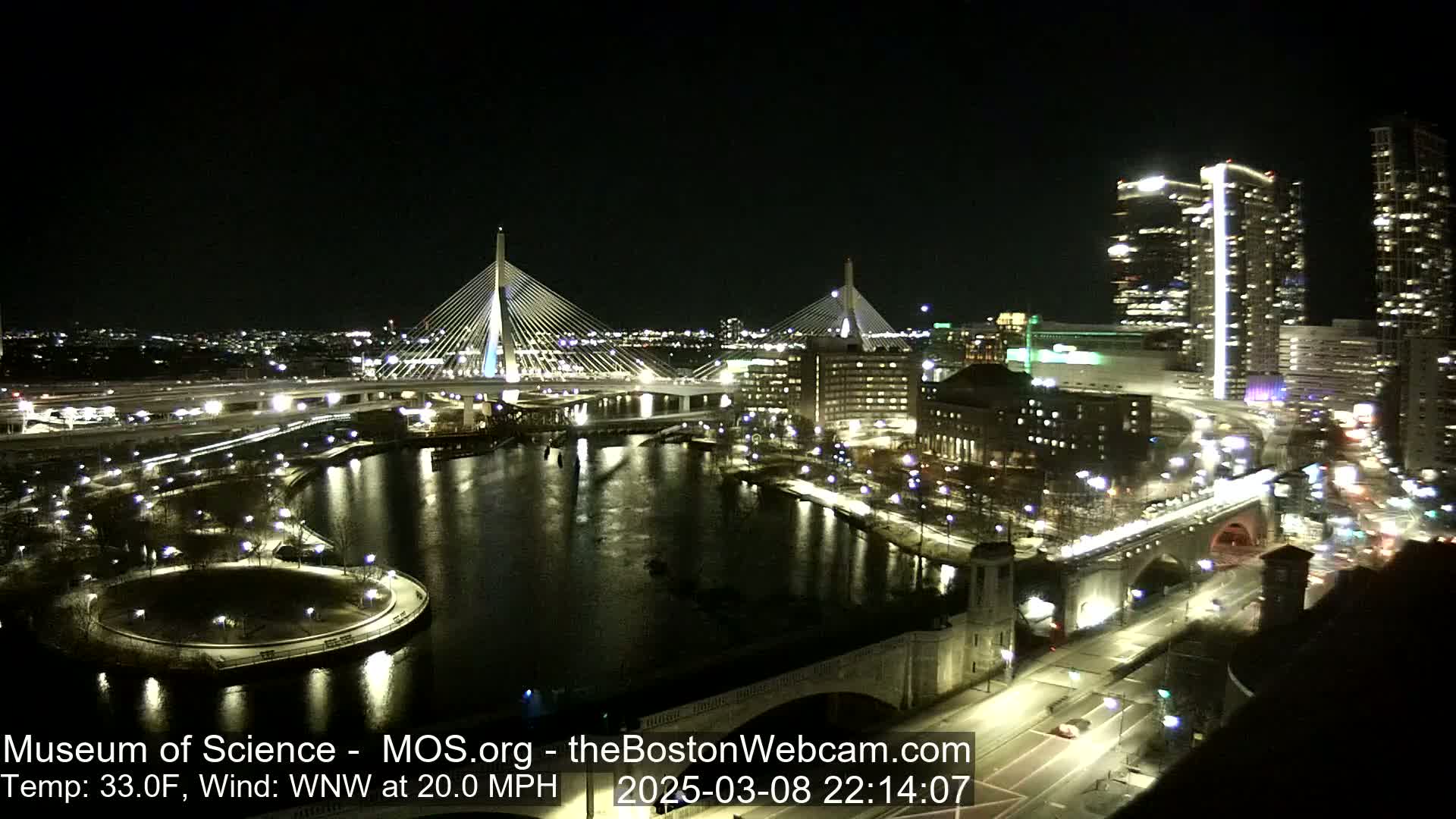 A nighttime view of a city waterfront with two illuminated cable-stayed bridges, a river, and city buildings, under a clear sky with a 20 mph wind.