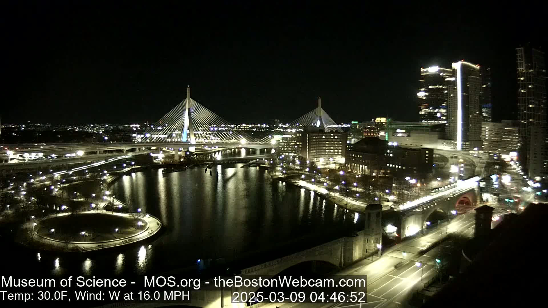 A nighttime view of a city waterfront with two cable-stayed bridges, illuminated buildings, and roadways, under a clear, windy sky with a temperature of 30°F.