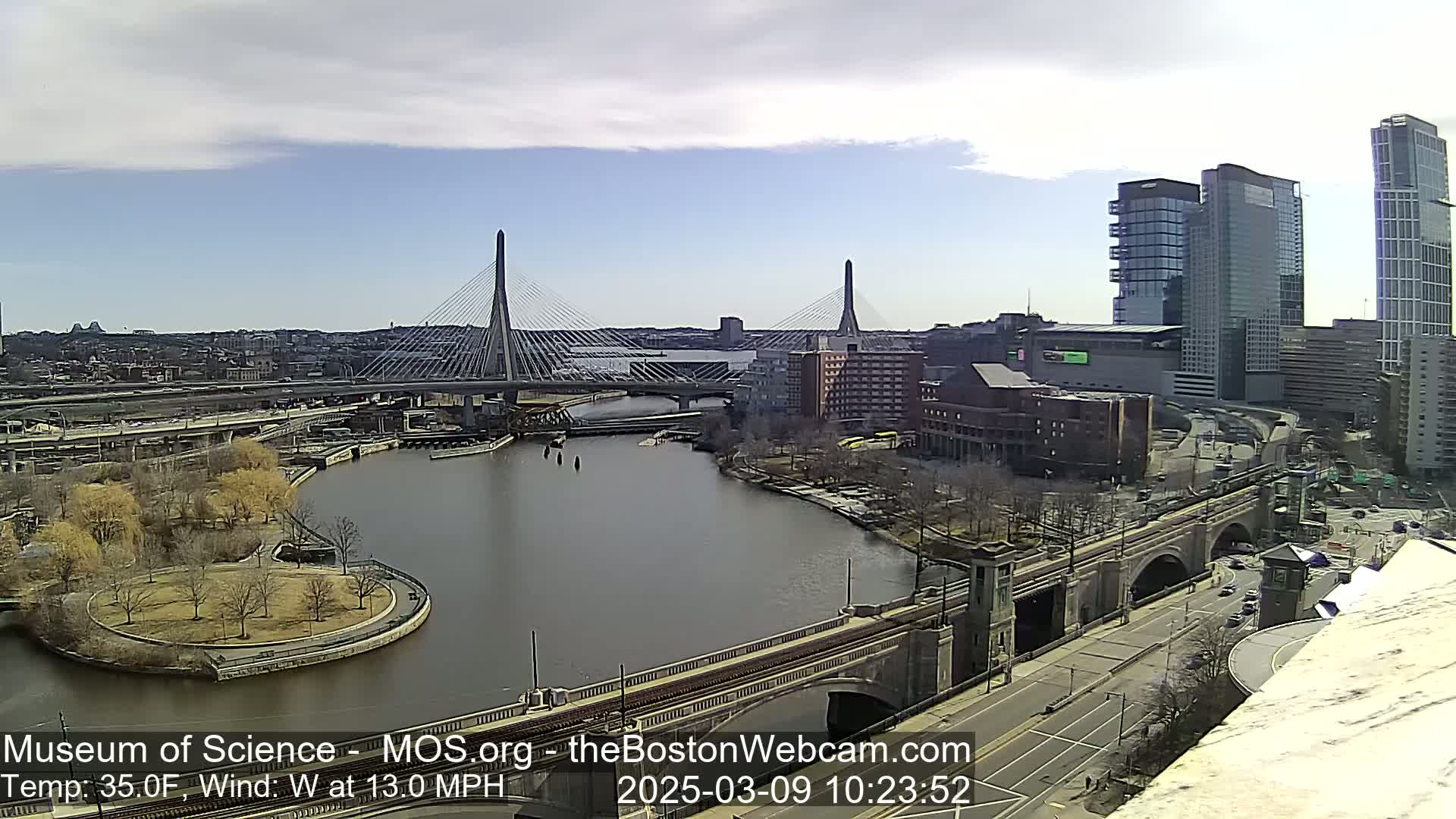 A city river scene on a partly cloudy day shows two cable-stayed bridges, a park, several buildings, and a roadway.