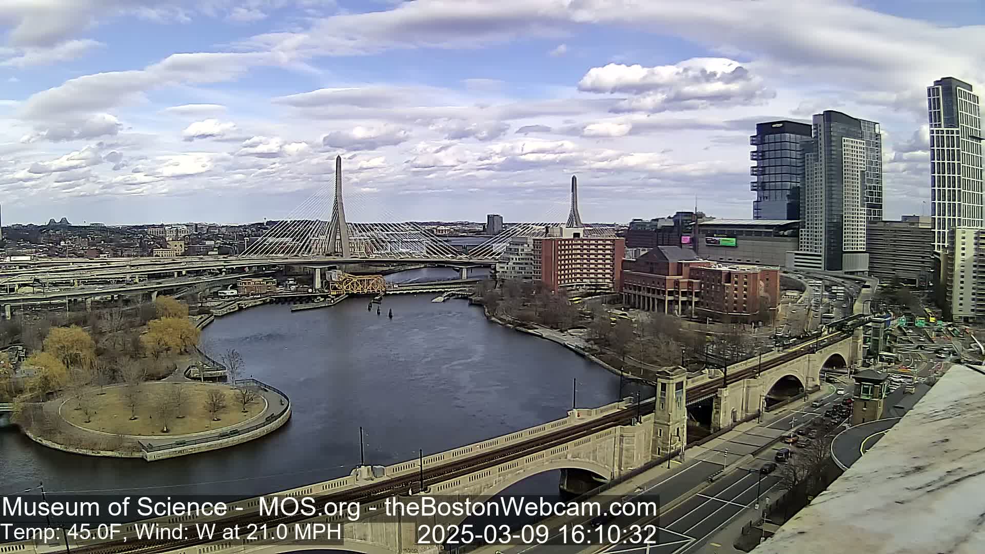 An aerial view of a city with two cable-stayed bridges spanning a river under partly cloudy skies, with a strong west wind.