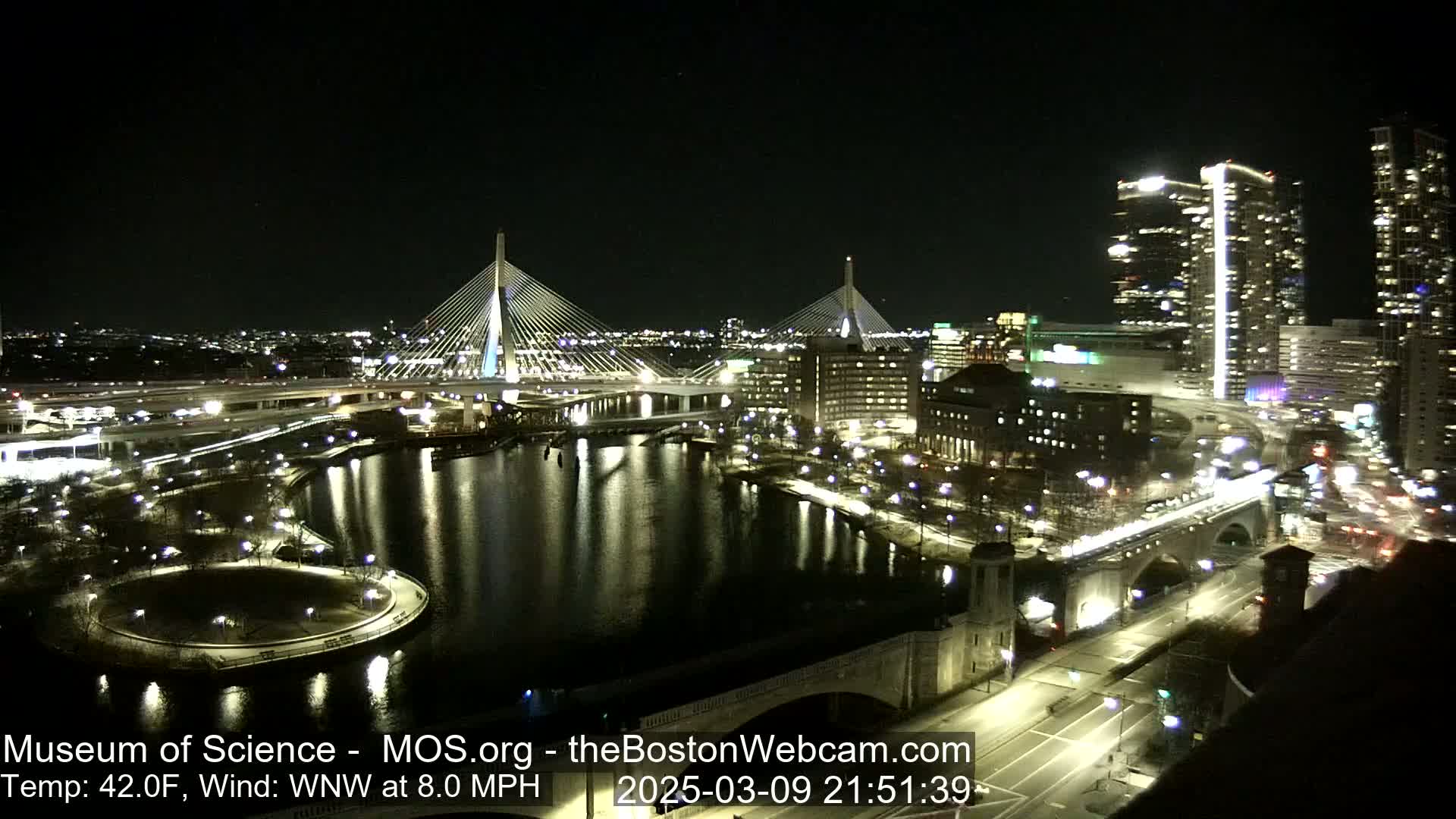 A nighttime view of a city with two illuminated cable-stayed bridges spanning a waterway, surrounded by city lights and buildings under a clear sky.