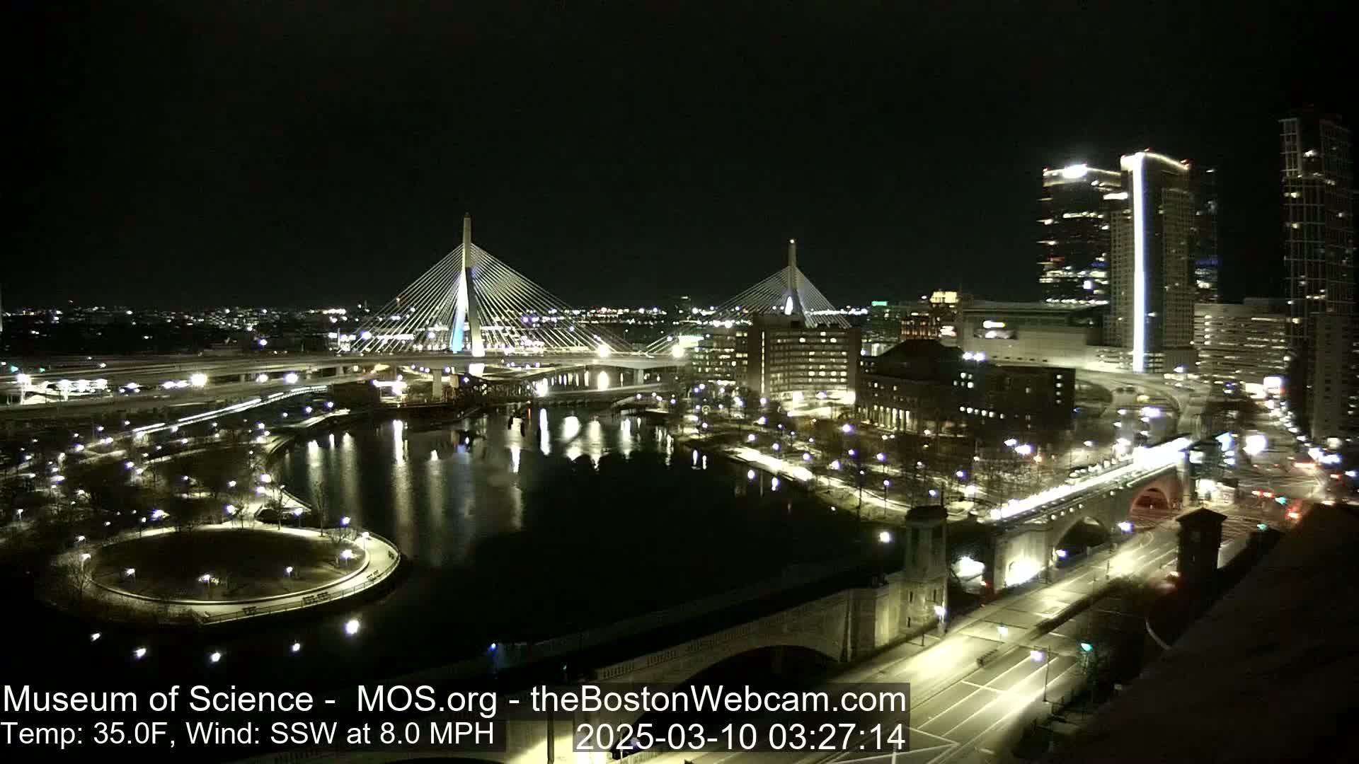 A nighttime cityscape featuring two illuminated cable-stayed bridges over a dark river, with city lights reflecting on the water under a clear sky and a slight south-southwesterly wind.