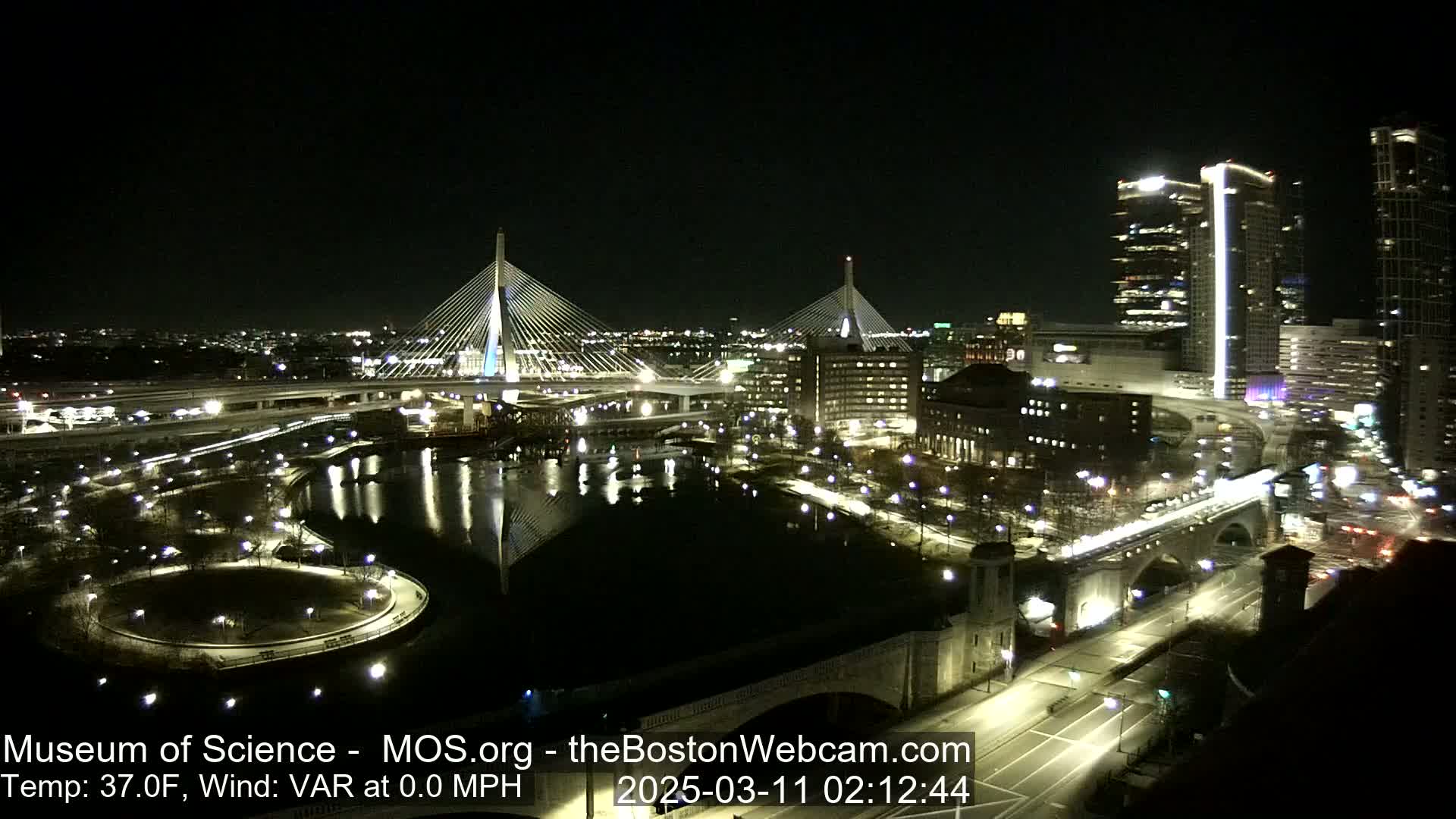 A nighttime view of two illuminated cable-stayed bridges spanning a dark river, with city lights and buildings visible in the background under a clear sky.
