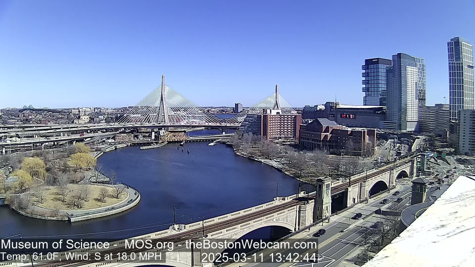 A sunny cityscape with two cable-stayed bridges spanning a river, under a clear blue sky, and a 18 mph south wind.