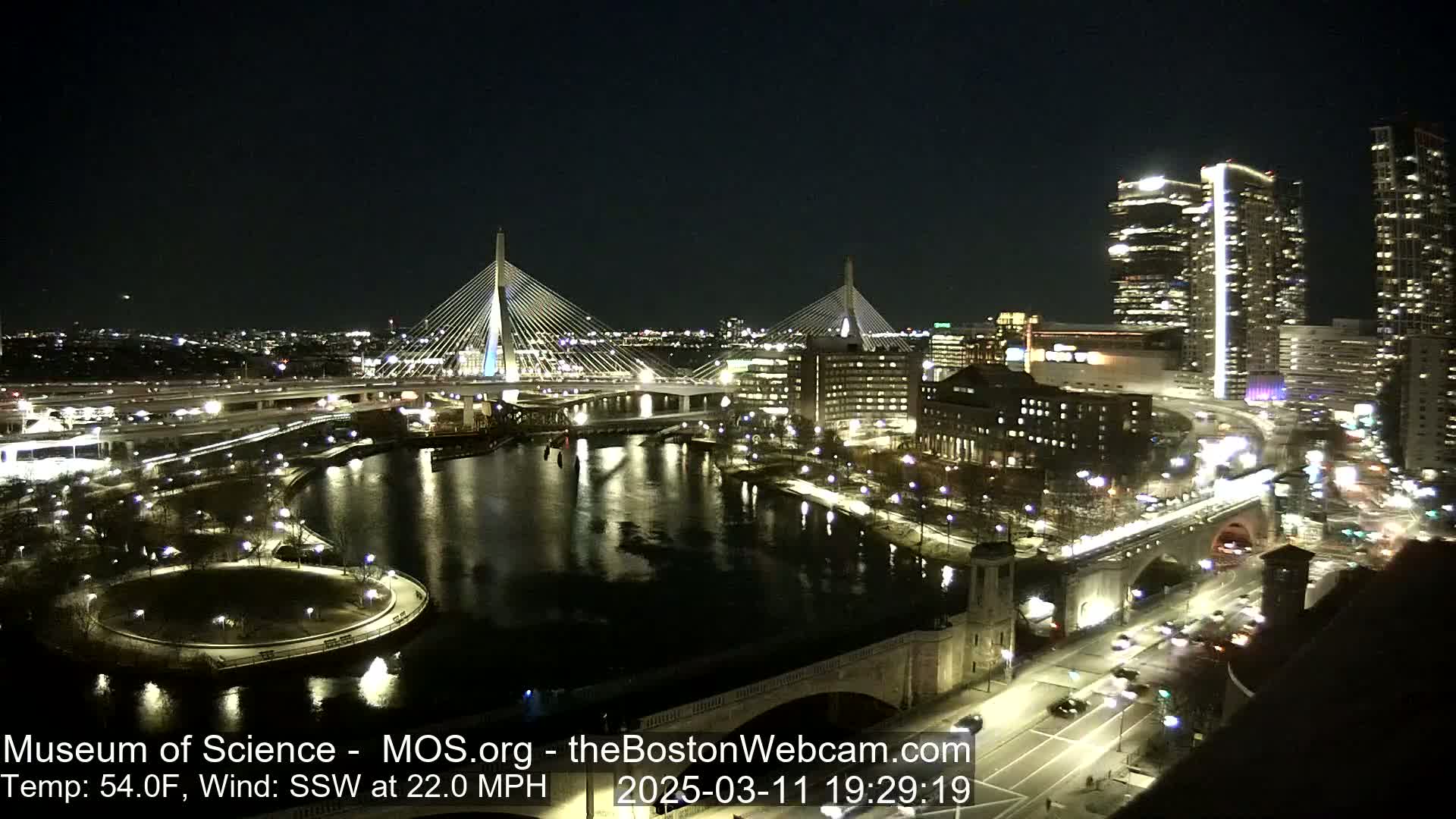 A nighttime, illuminated cityscape features two cable-stayed bridges spanning a dark river, with city lights reflecting in the water under a clear, dark sky, and a strong south-southwesterly wind.