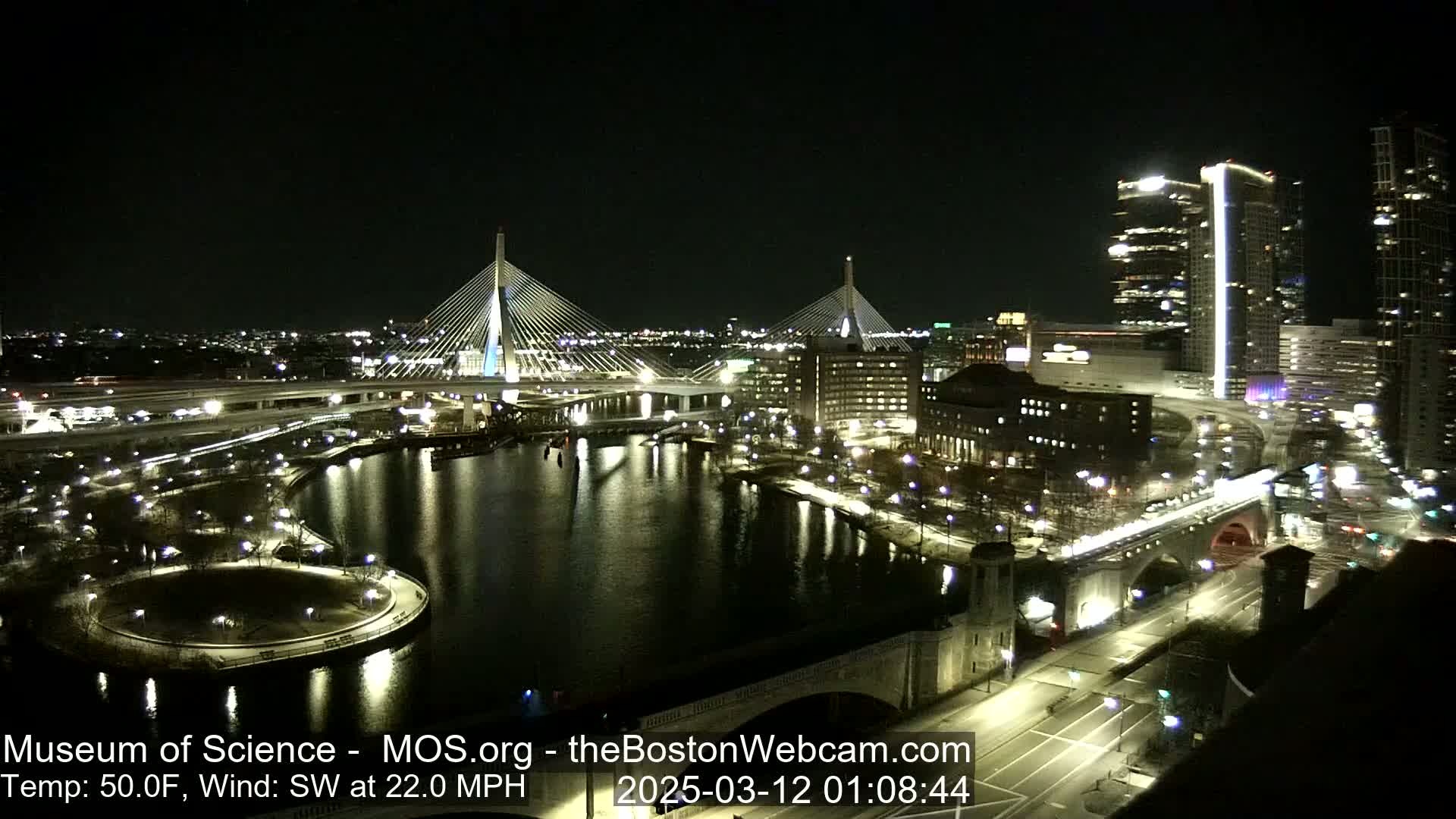 A nighttime view of a city waterfront with two cable-stayed bridges, illuminated buildings, and roadways, under a clear sky and with a southwest wind at 22 mph.